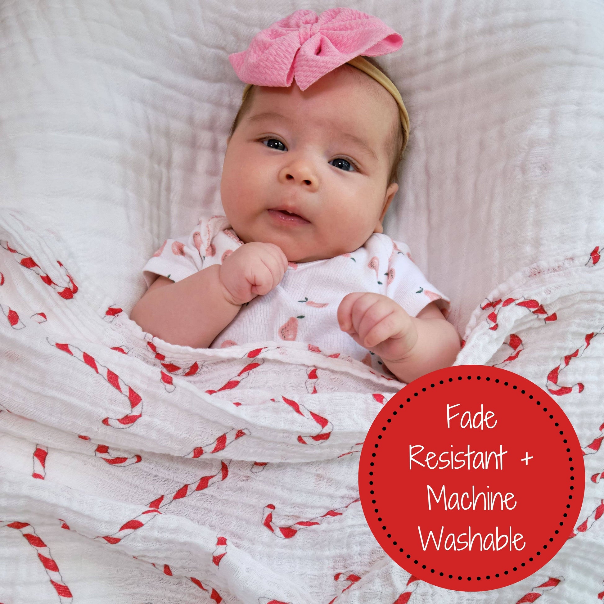 Baby wrapped in a red and white patterned blanket with a pink bow, on a white textured surface.
