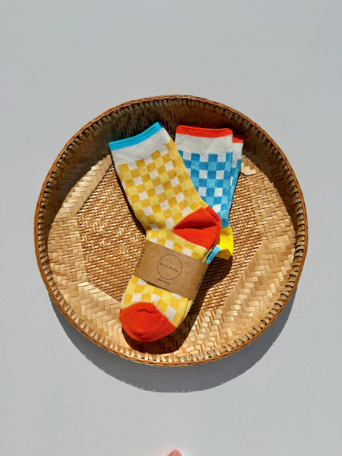 A pair of colorful patterned socks displayed inside a round woven straw basket on a light gray background. The socks feature yellow and white checkerboard patterns on one leg, blue and white checkerboard patterns on the other, with bright orange toes and heels, and a brown paper band around the ankle.