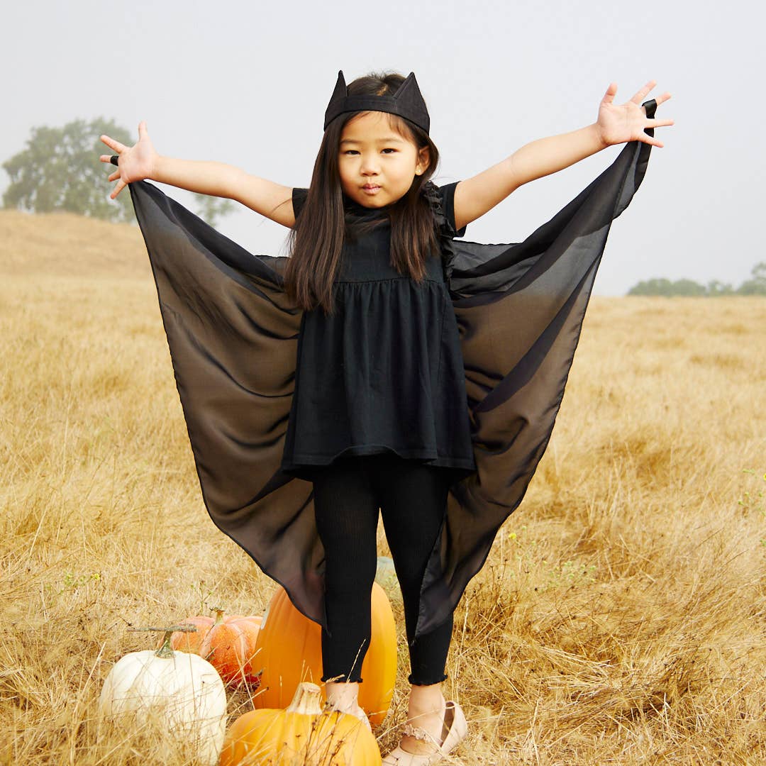 A young girl wearing a black bat costume stands in a dry grass field with pumpkins at her feet, holding out her black sheer cape-like wingspan.