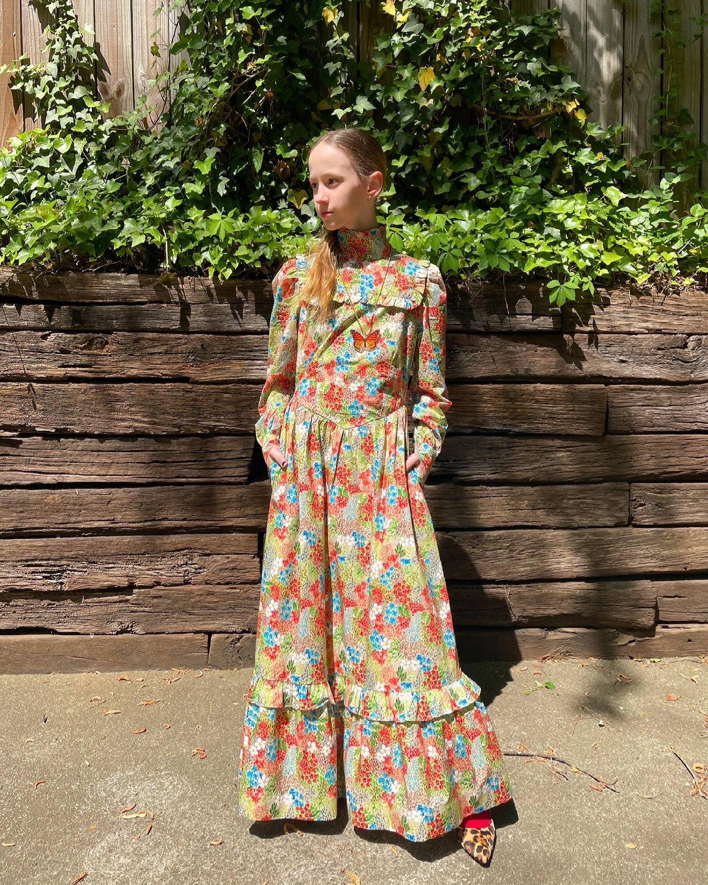 Woman in a floral dress standing against a wooden wall with greenery.