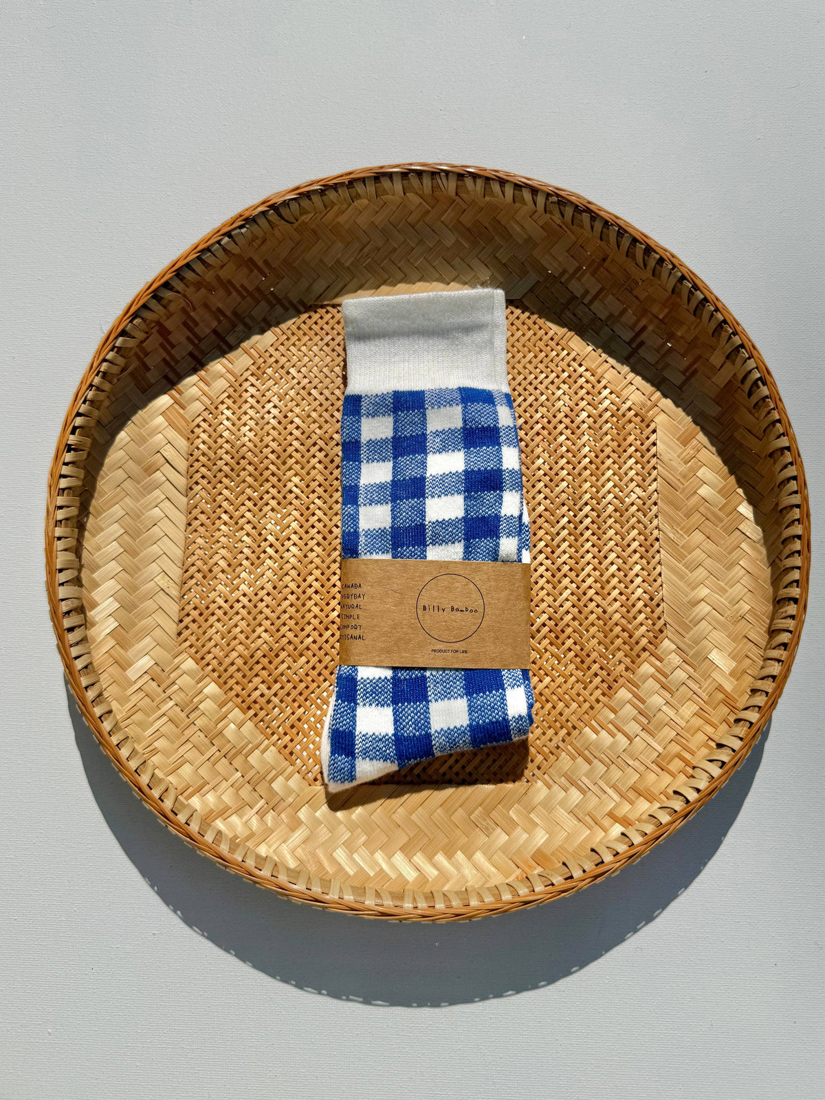 A pair of blue and white gingham checkered socks folded and wrapped in a kraft paper band, placed inside a round woven straw tray on a light gray background.