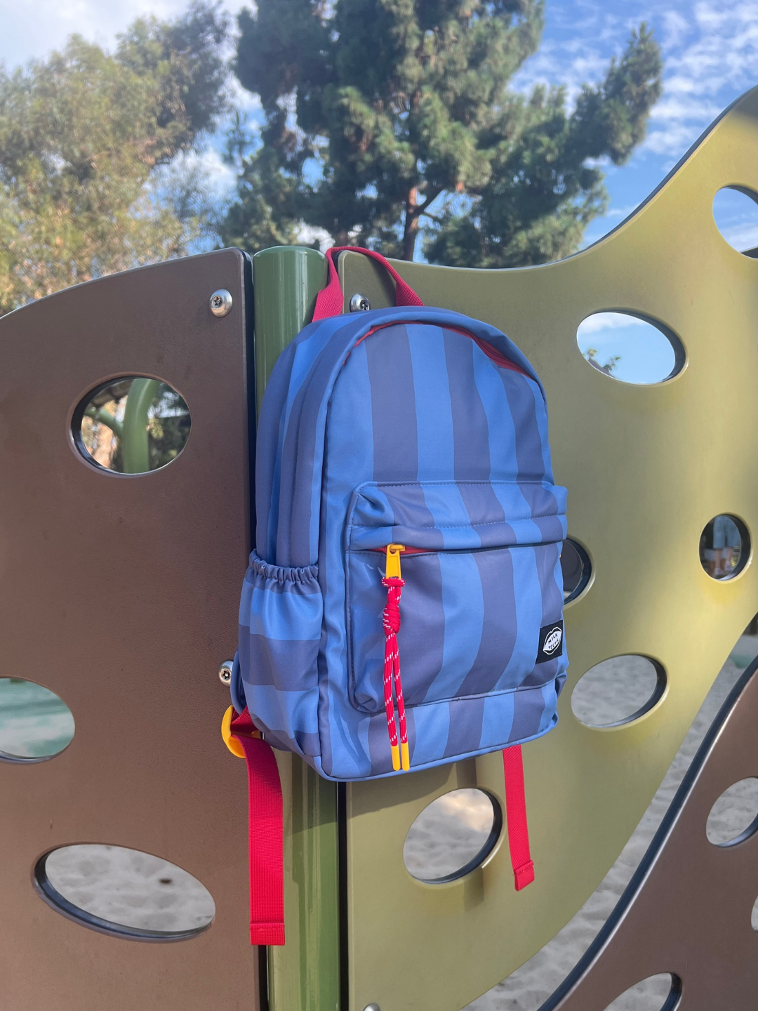 A blue and purple striped backpack with red straps and a red zipper pull hanging on a playground metal structure outdoors.