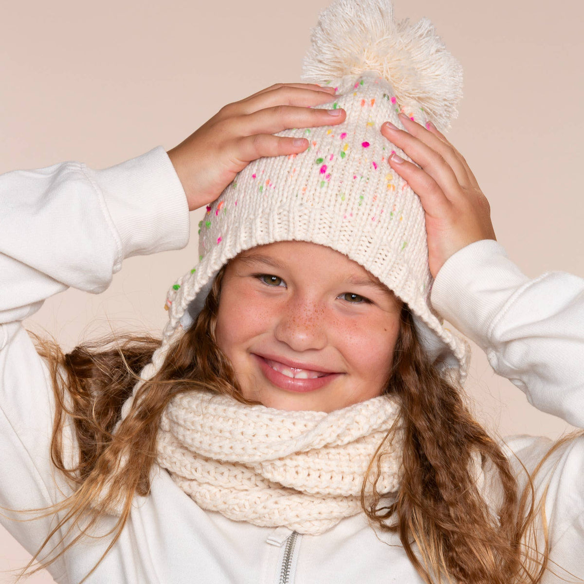 A young girl smiling while wearing a cream knit pom-pom beanie with colorful speckles and a matching cream knit neck warmer, posed against a solid pale pink background.