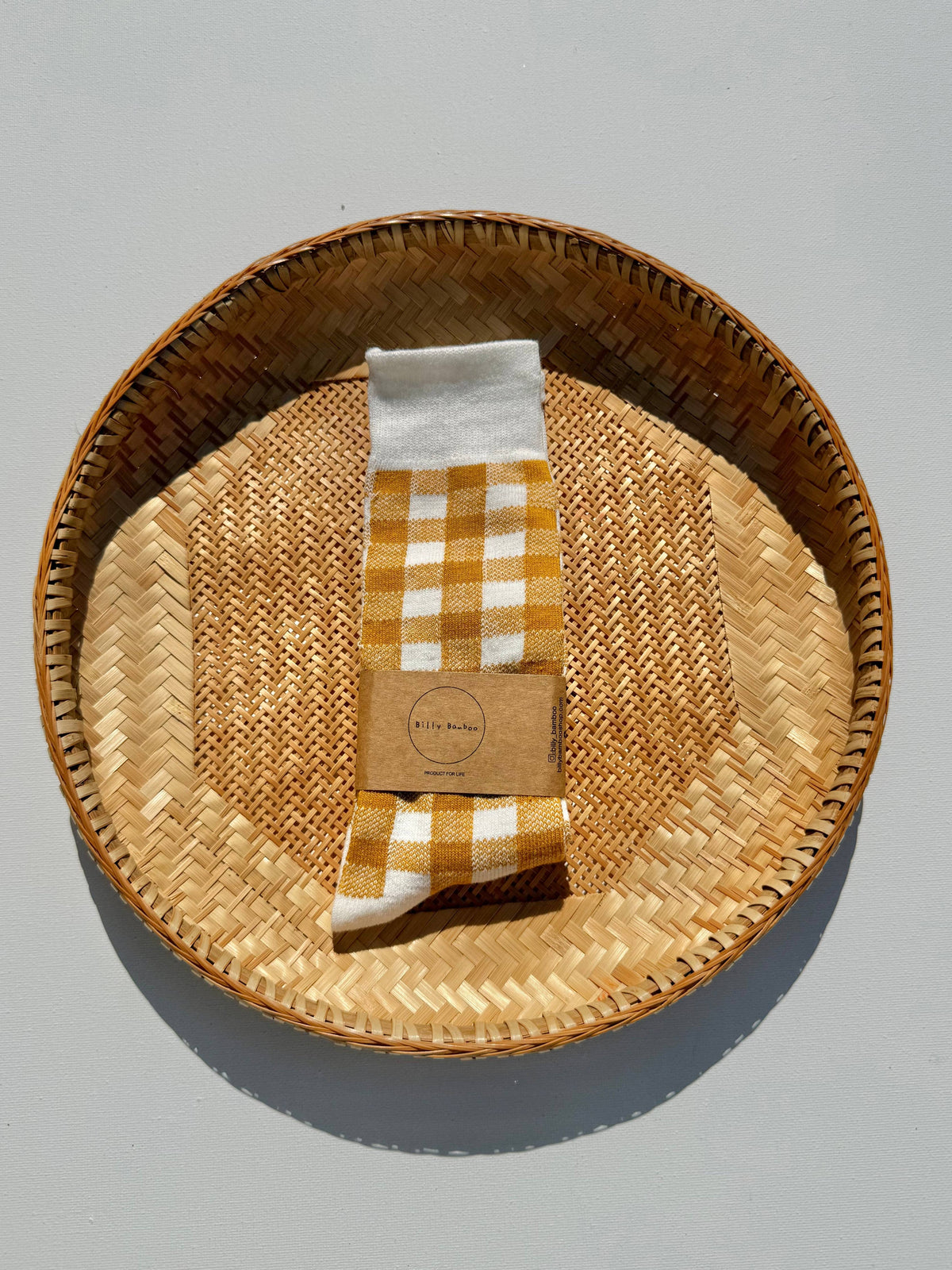 A pair of yellow and white gingham check socks with a brown kraft paper band, presented inside a round woven rattan tray on a neutral gray background.