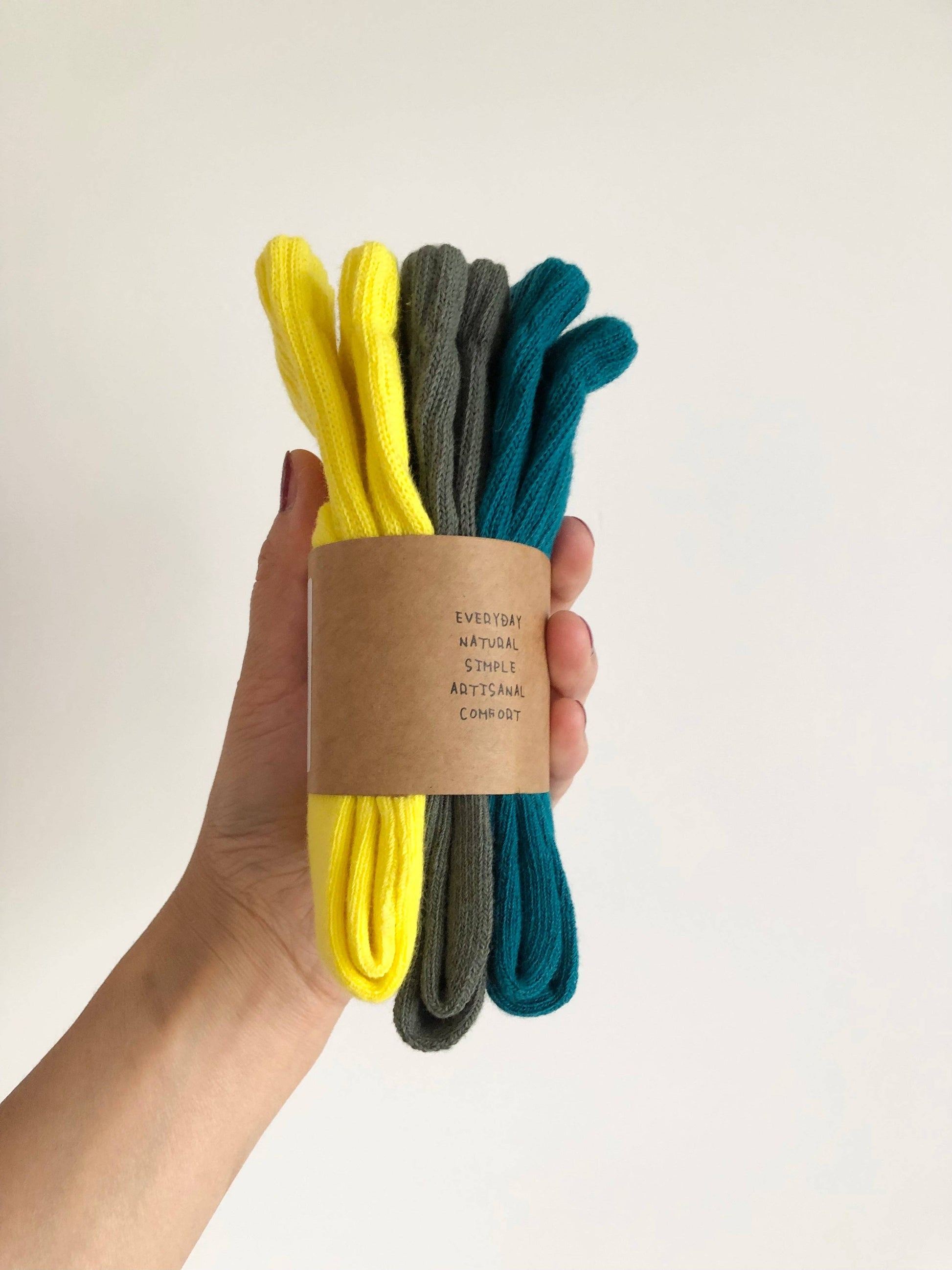 Hand holding a bundle of three pairs of socks in yellow, gray, and blue with a cardboard sleeve on a light background.