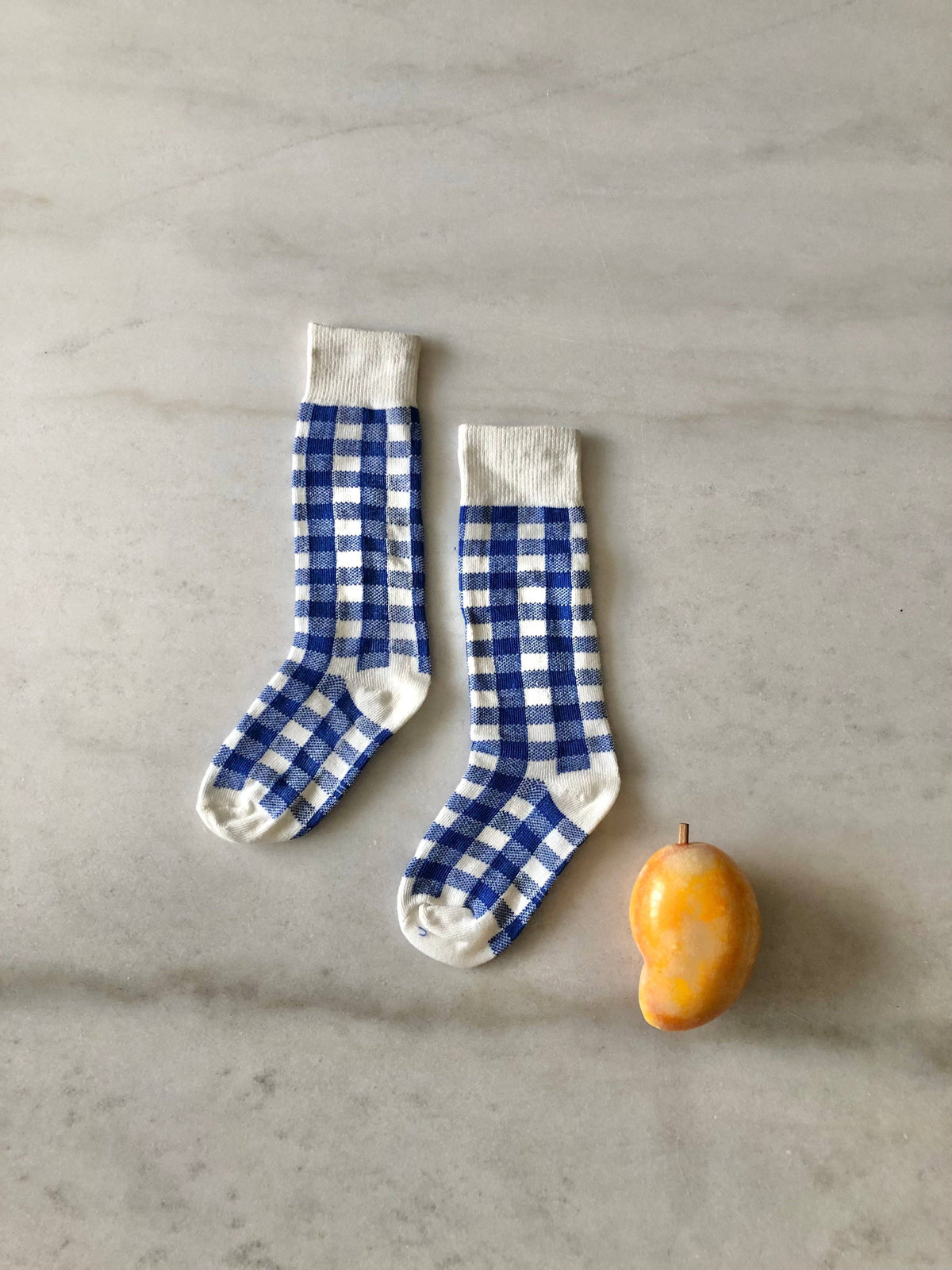 A pair of blue and white gingham check socks laid flat on a light gray marble surface next to a small yellow-orange mango.
