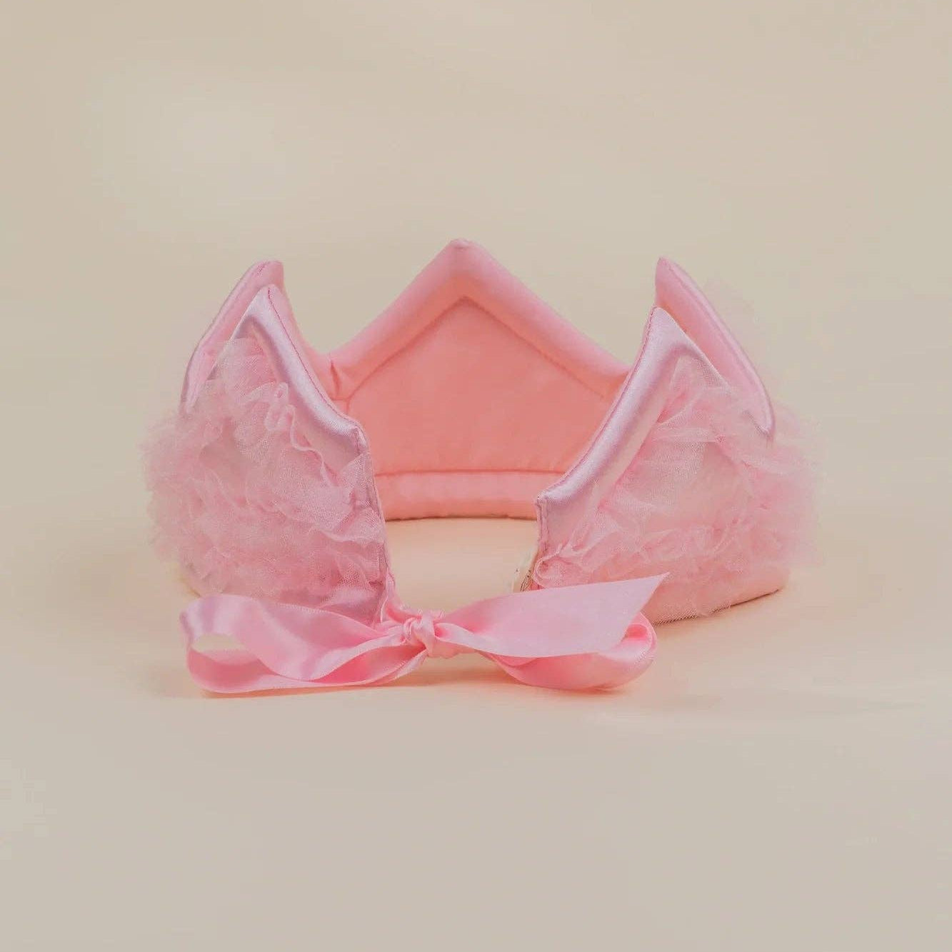 A pink plush cat-ear headband with fluffy tulle details and a matching pink satin bow, displayed on a solid light beige background.