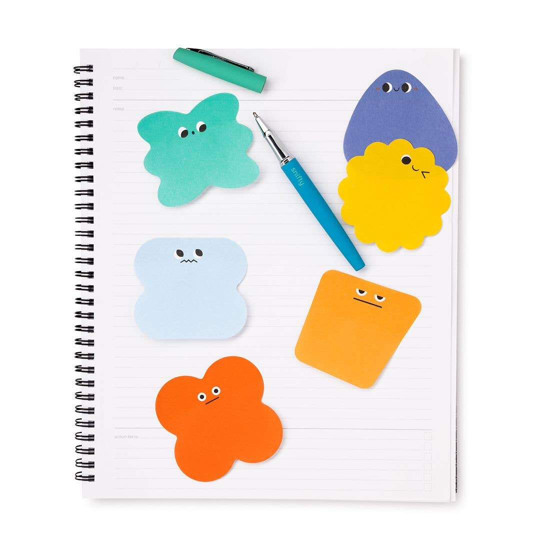 A spiral-bound lined notebook on a white background, decorated with colorful cartoon-shaped sticky notes in teal, blue, yellow, orange, and light blue, along with a teal pen and a blue pen.