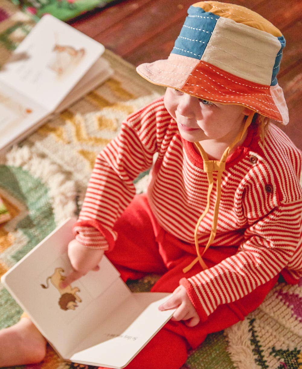 Child wearing a colorful hat and red outfit, sitting on a patterned rug with a book.