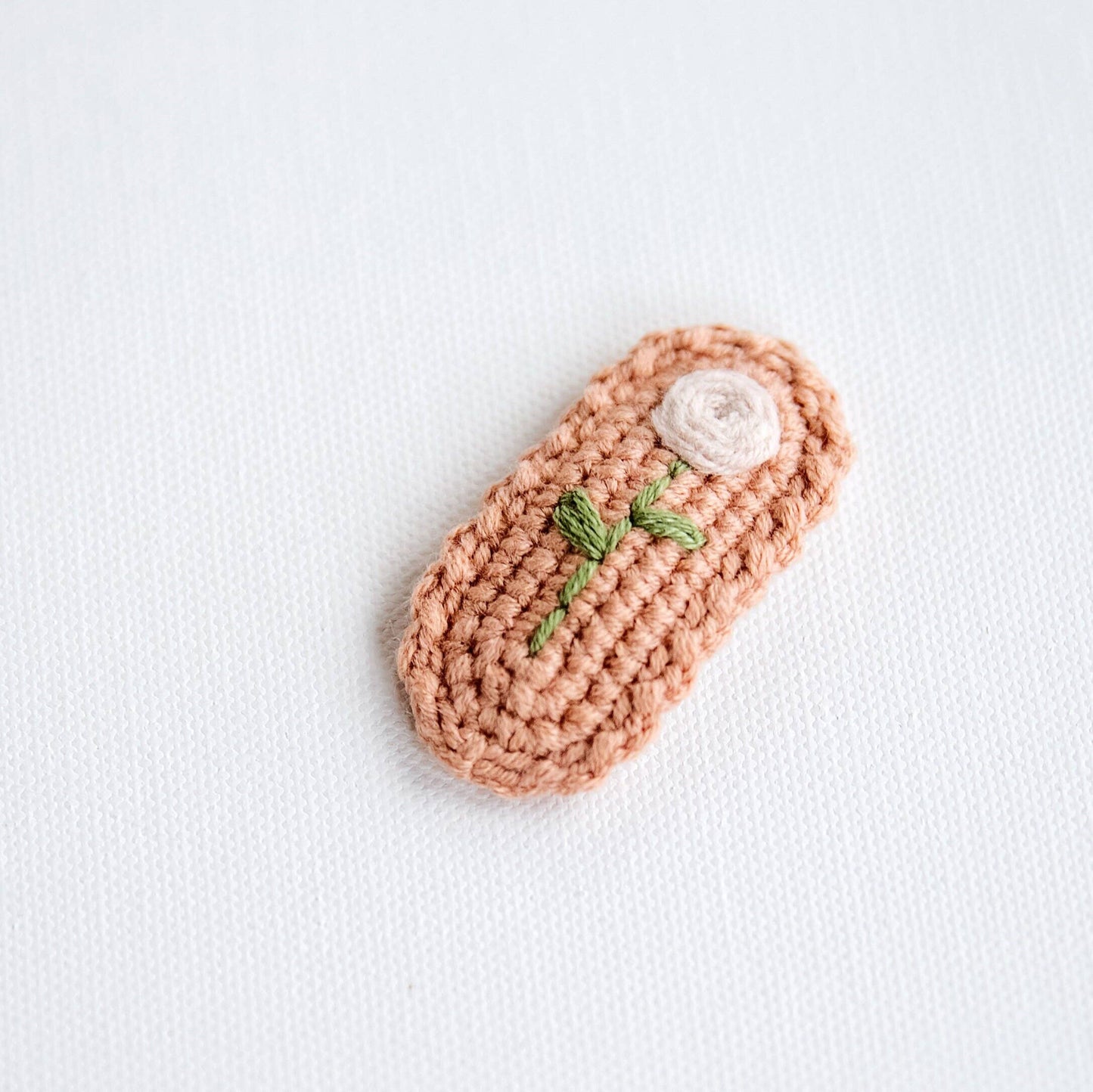 Crocheted peach-shaped object with a white flower and green leaves on a light gray background