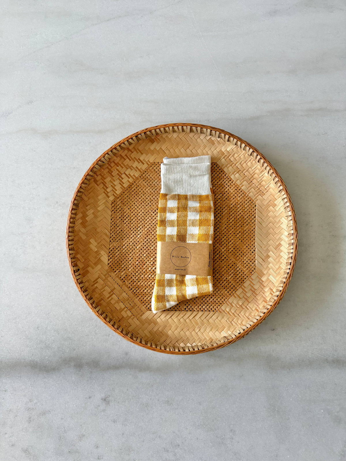 A folded yellow and white gingham socks with a kraft paper band label, placed on top of a round woven rattan tray over a light gray marble surface.