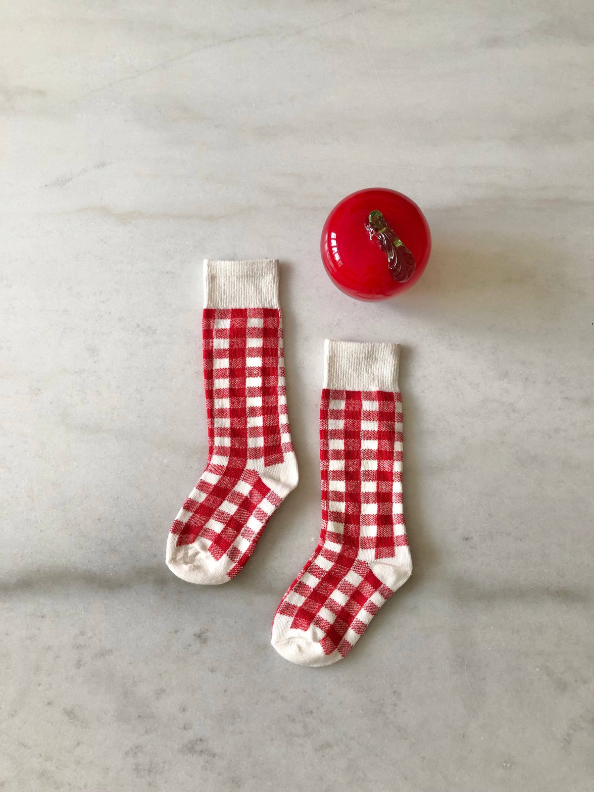 A pair of red and white gingham check baby socks with white ribbed cuffs, displayed on a light gray marble surface next to a small red decorative ball.