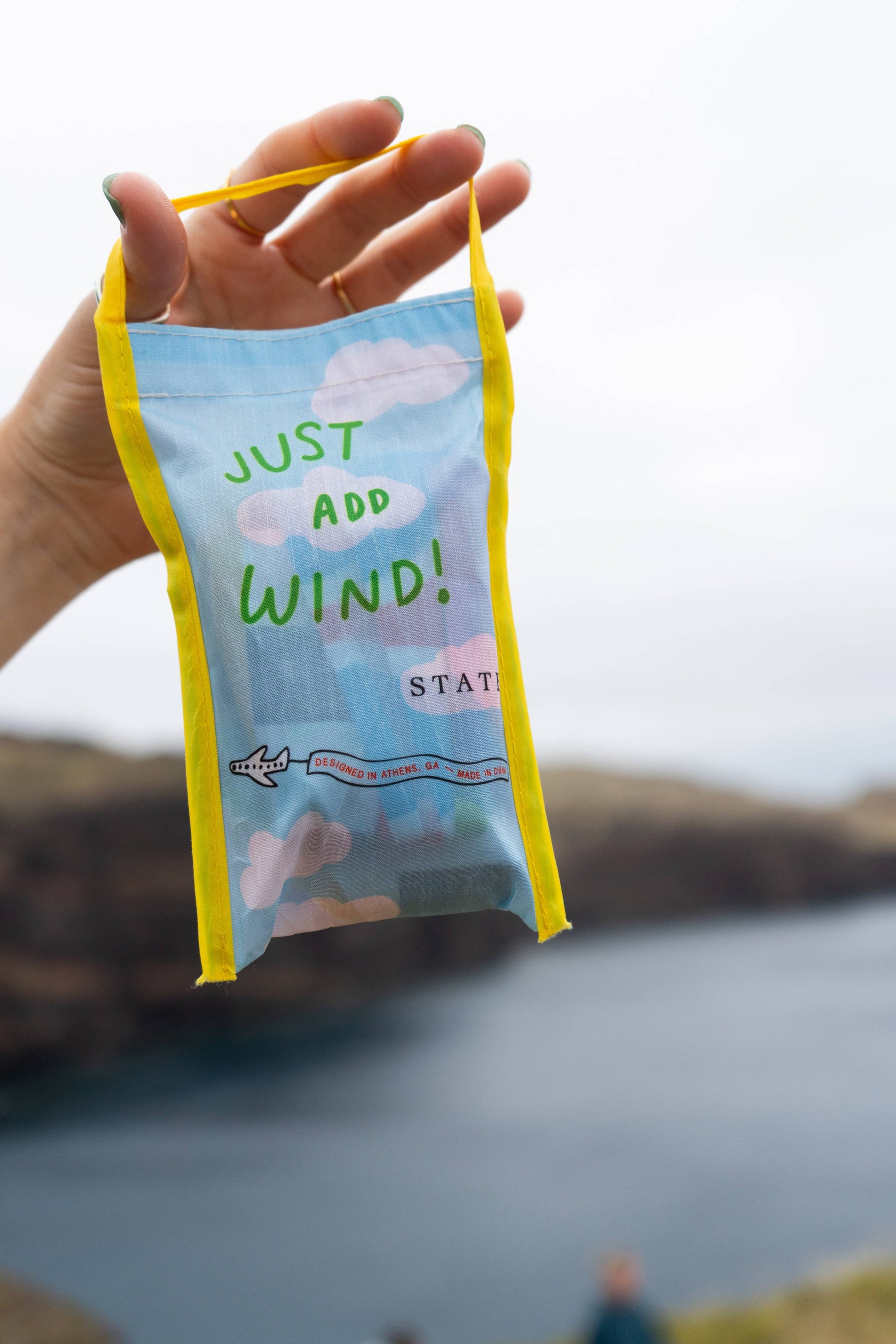 Hand holding a small blue bag with text 'Just Add Wind' against a blurred natural background