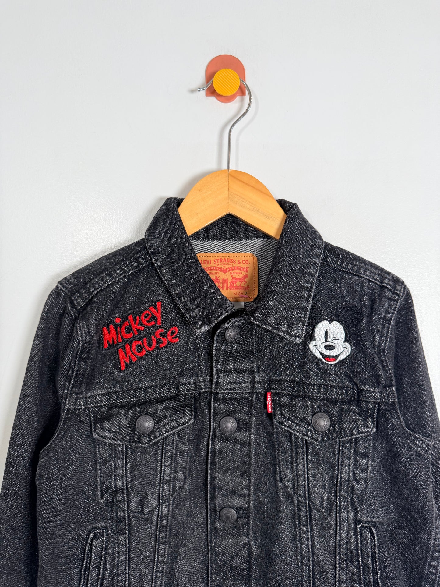 Black denim jacket with 'Mickey Mouse' embroidery on a wooden hanger against a white background