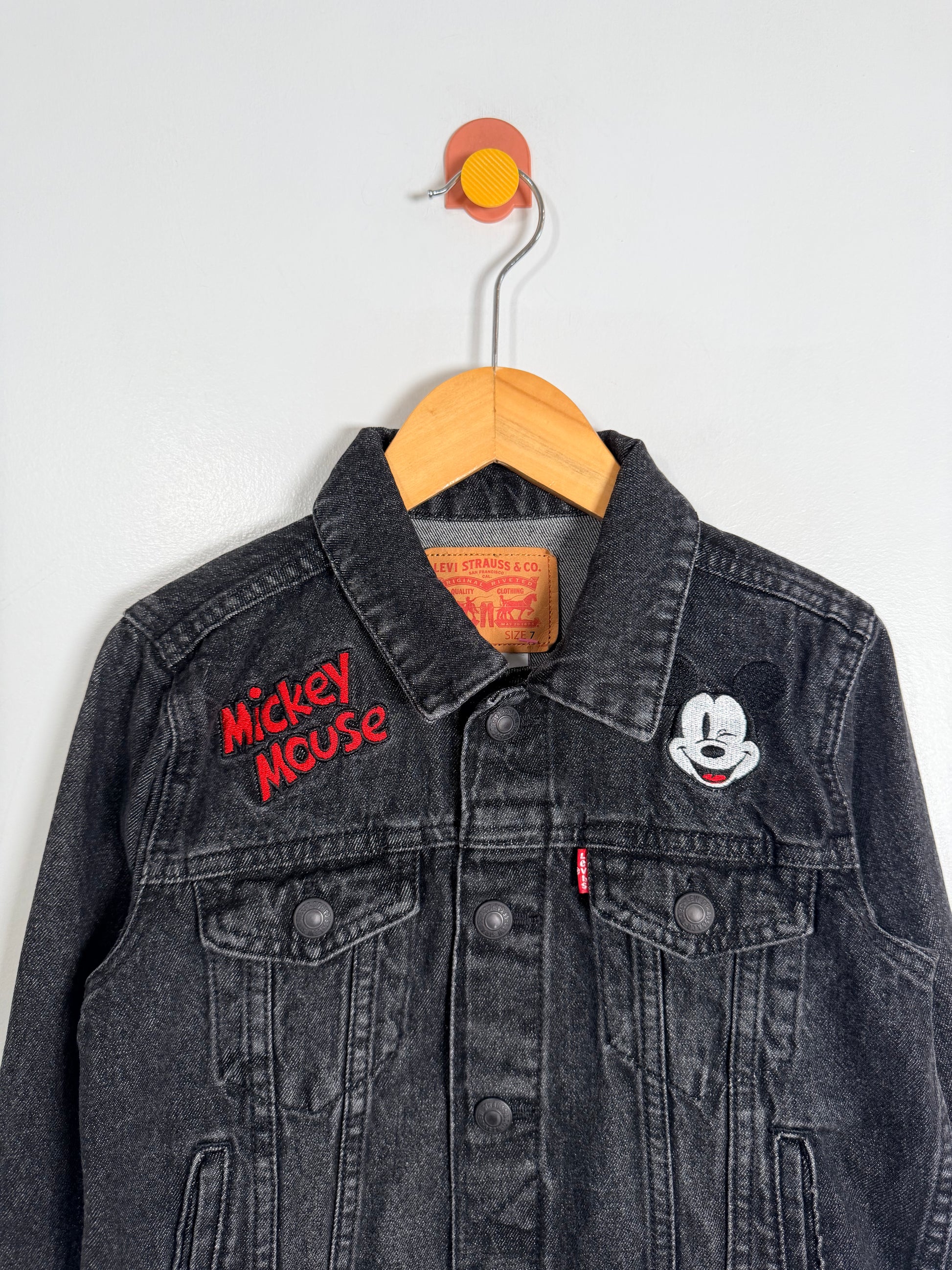 Black denim jacket with 'Mickey Mouse' embroidery on a wooden hanger against a white background