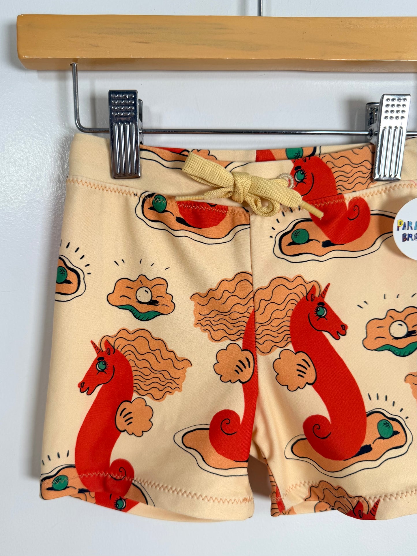 Beige swim horts with red horse design hanging on a wooden hanger.