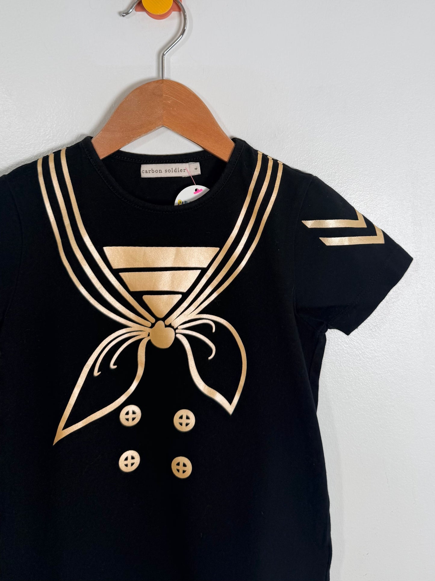 Black t-shirt with gold nautical design on a wooden hanger against a white background