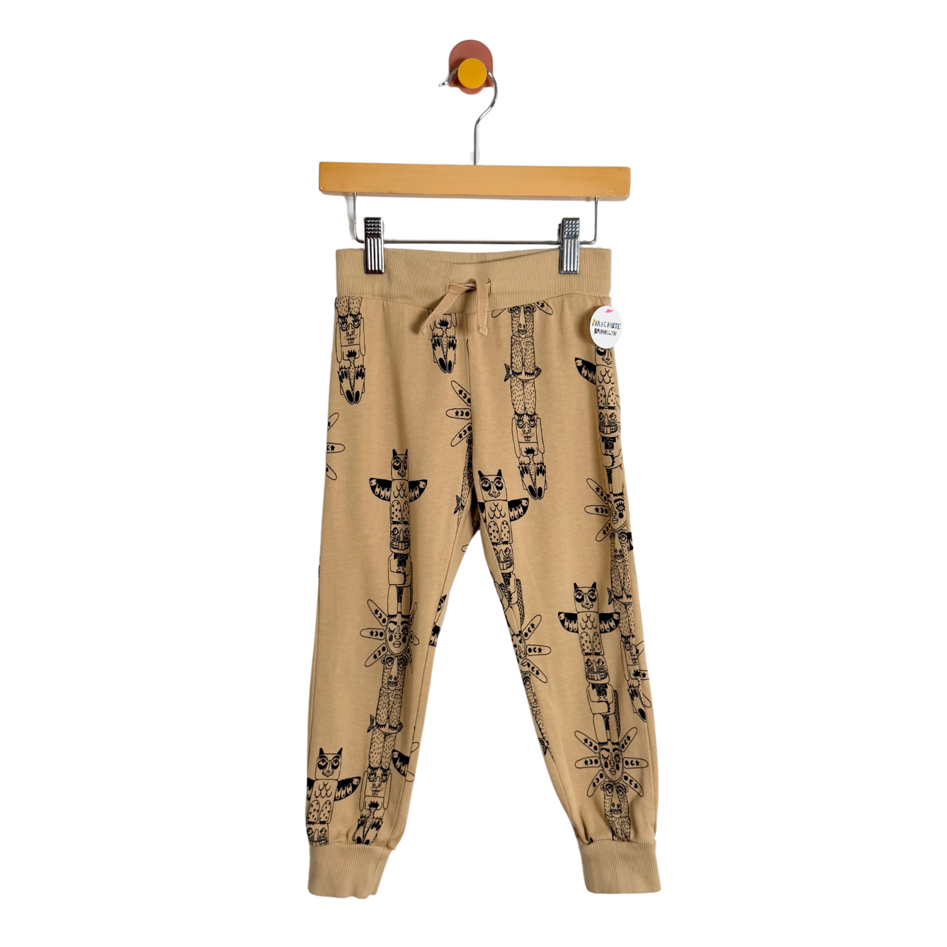 Beige sweatpants with black totem print on a hanger against a white background