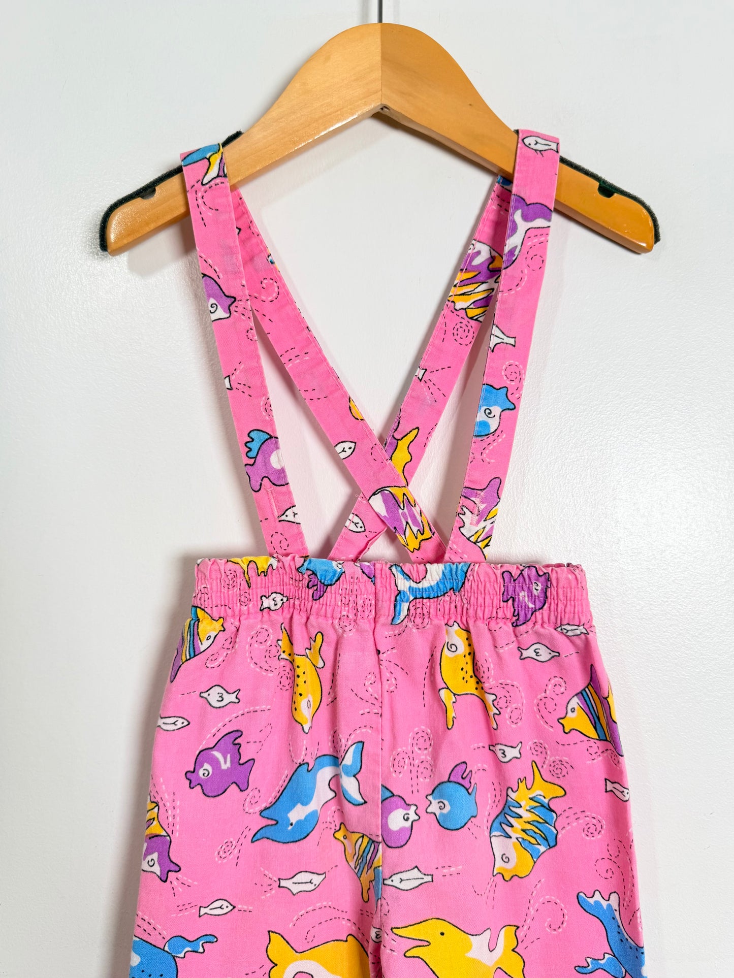 Pink children's overalls with colorful fish pattern on a hanger against a white background