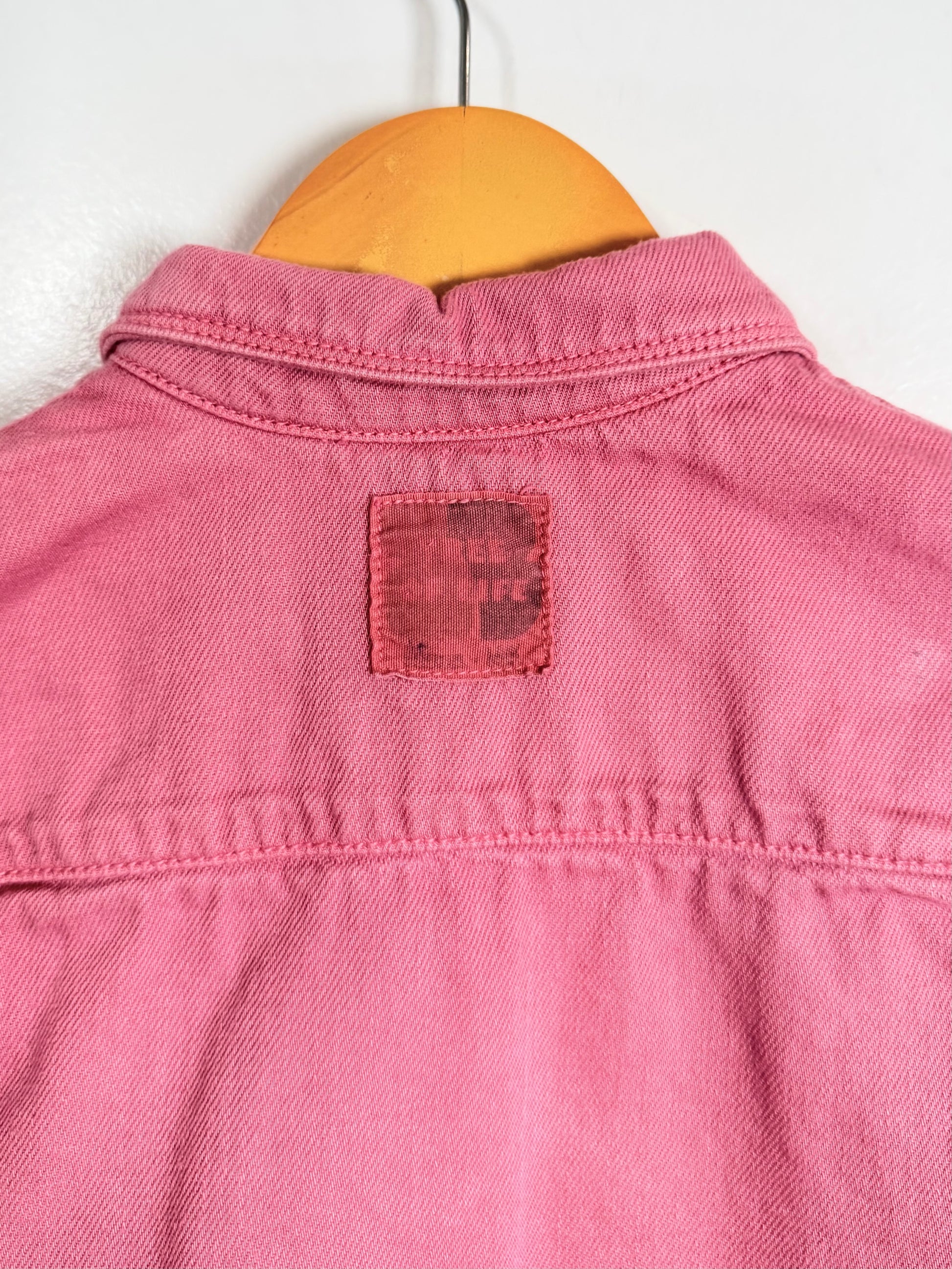 Pink denim shirt with a brand label on a wooden hanger against a white background
