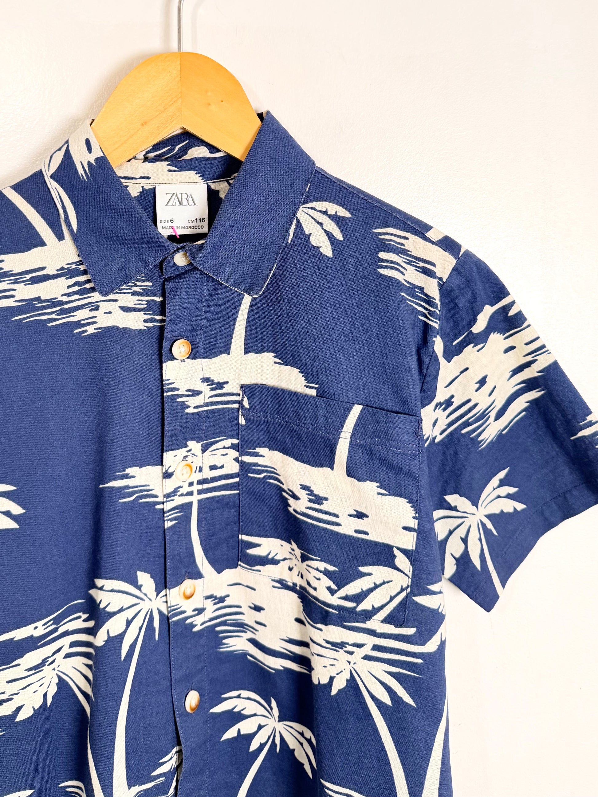 Blue shirt with white palm tree pattern on a hanger against a white background