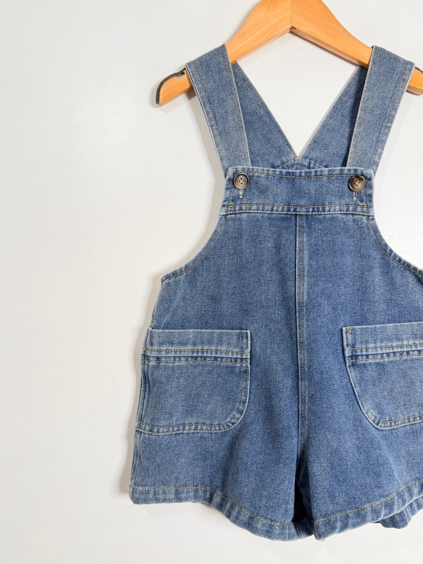 Blue denim overall shorts on a wooden hanger against a white background