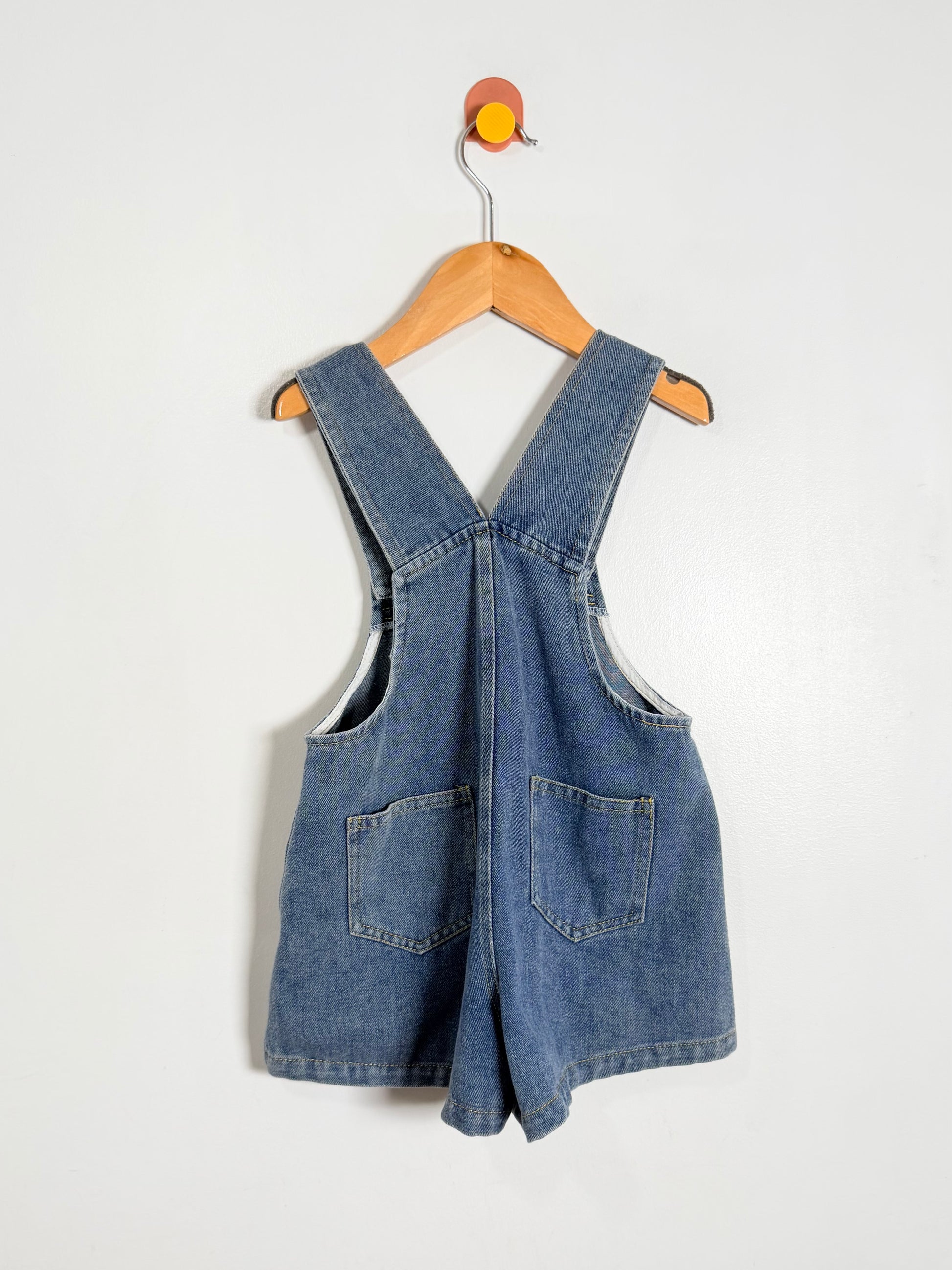 Blue denim overall dress on a wooden hanger against a white background