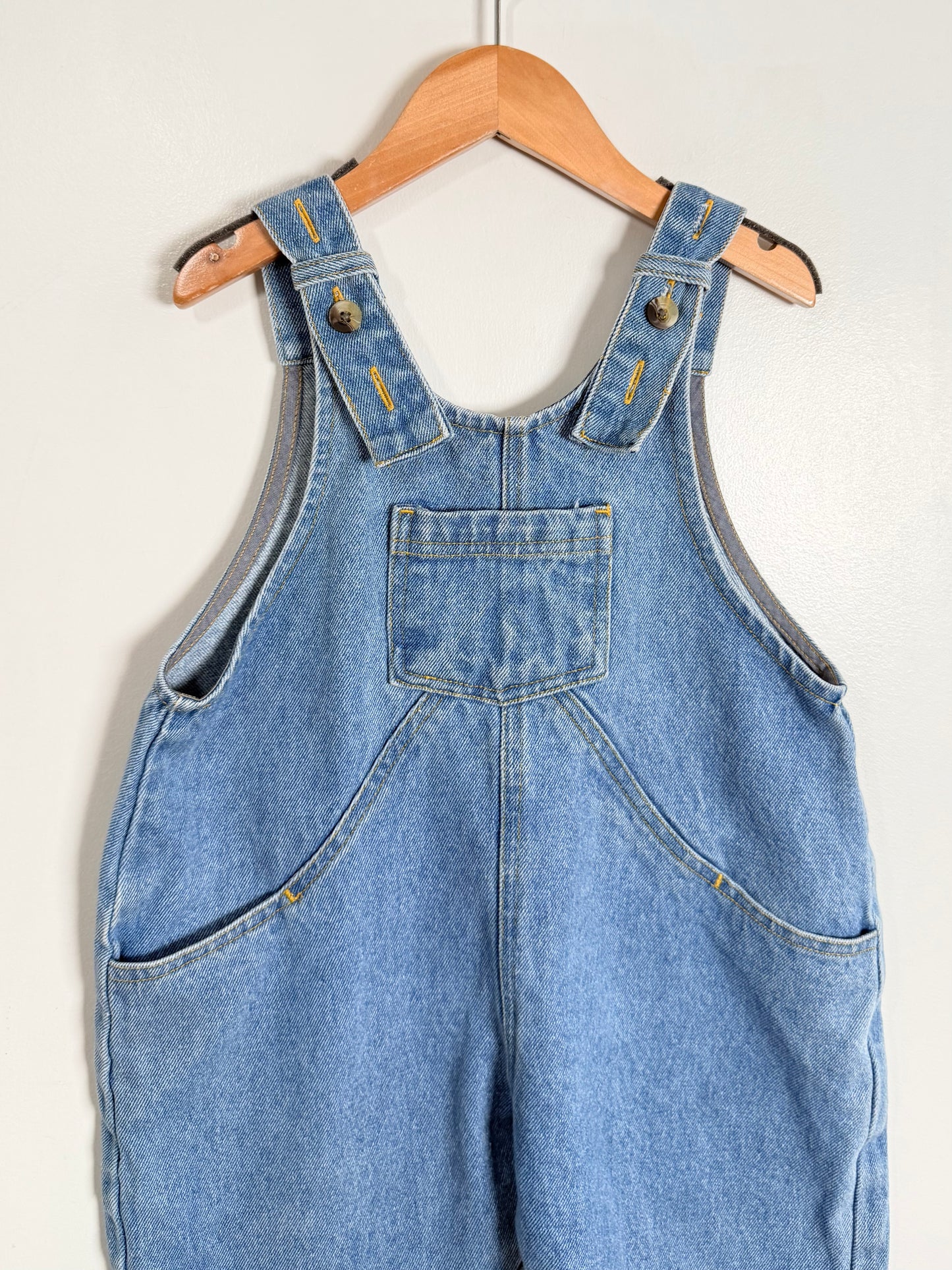 Blue denim overalls hanging on a wooden hanger against a white background