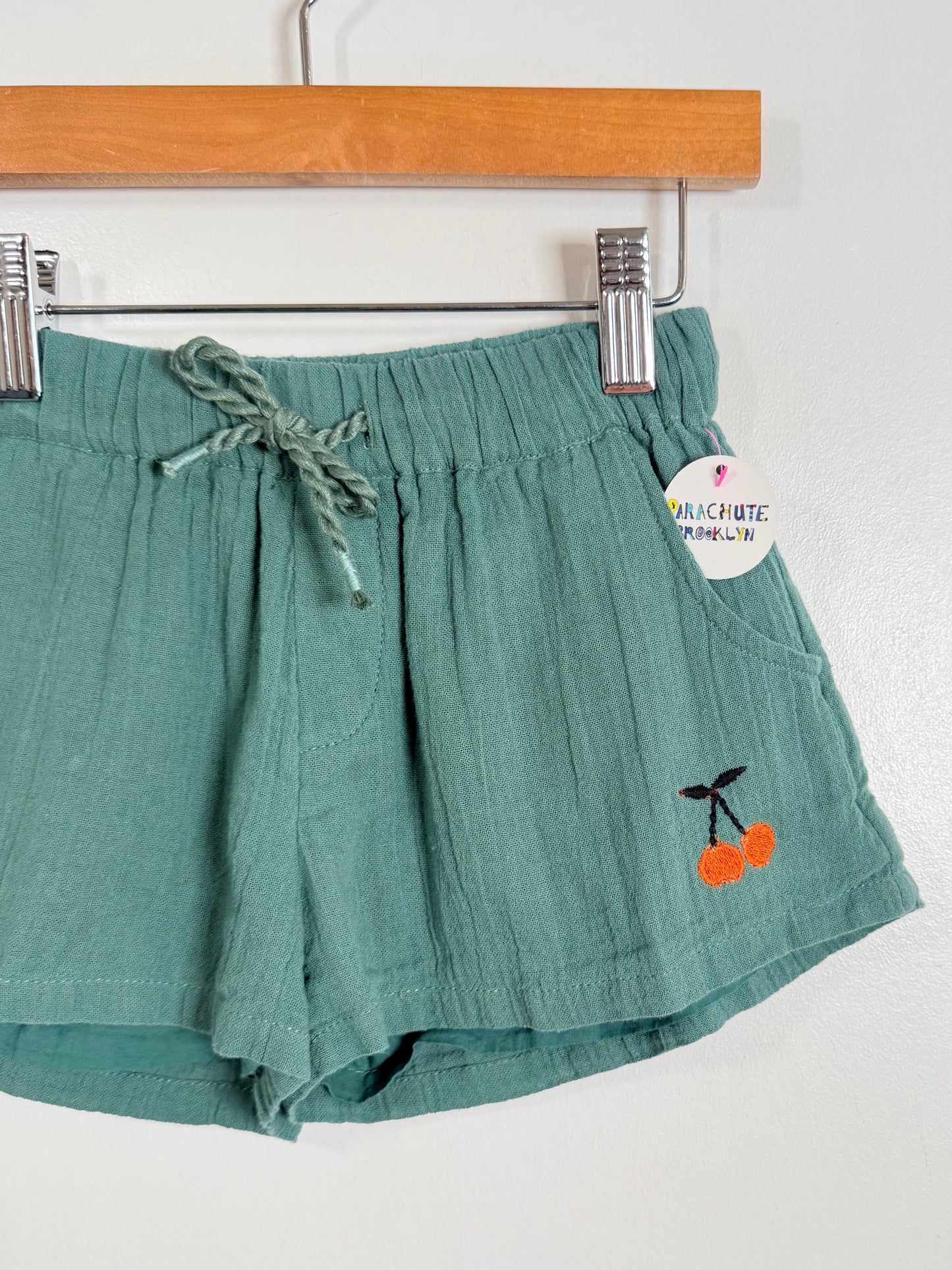 Green shorts with cherry embroidery on a hanger against a white background