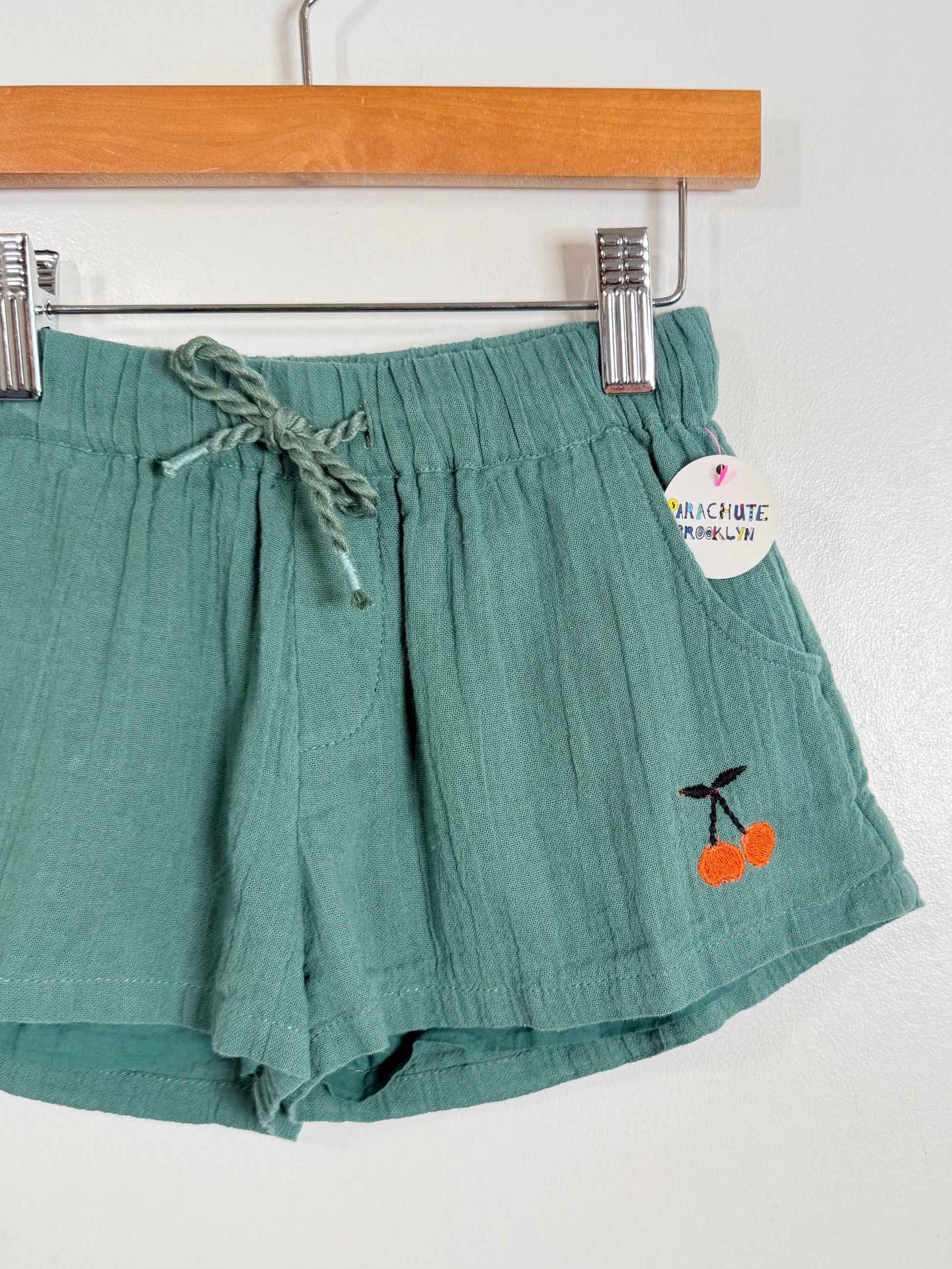 Green shorts with cherry embroidery on a hanger against a white background