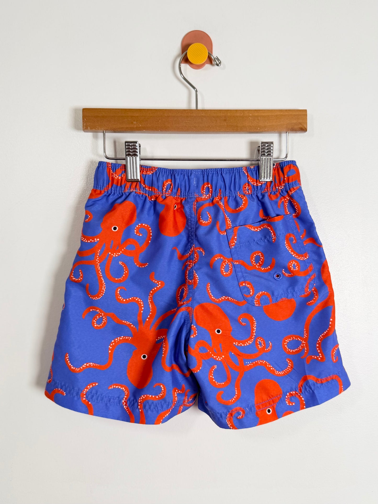 Blue swim shorts with orange octopus pattern on a hanger against a white background