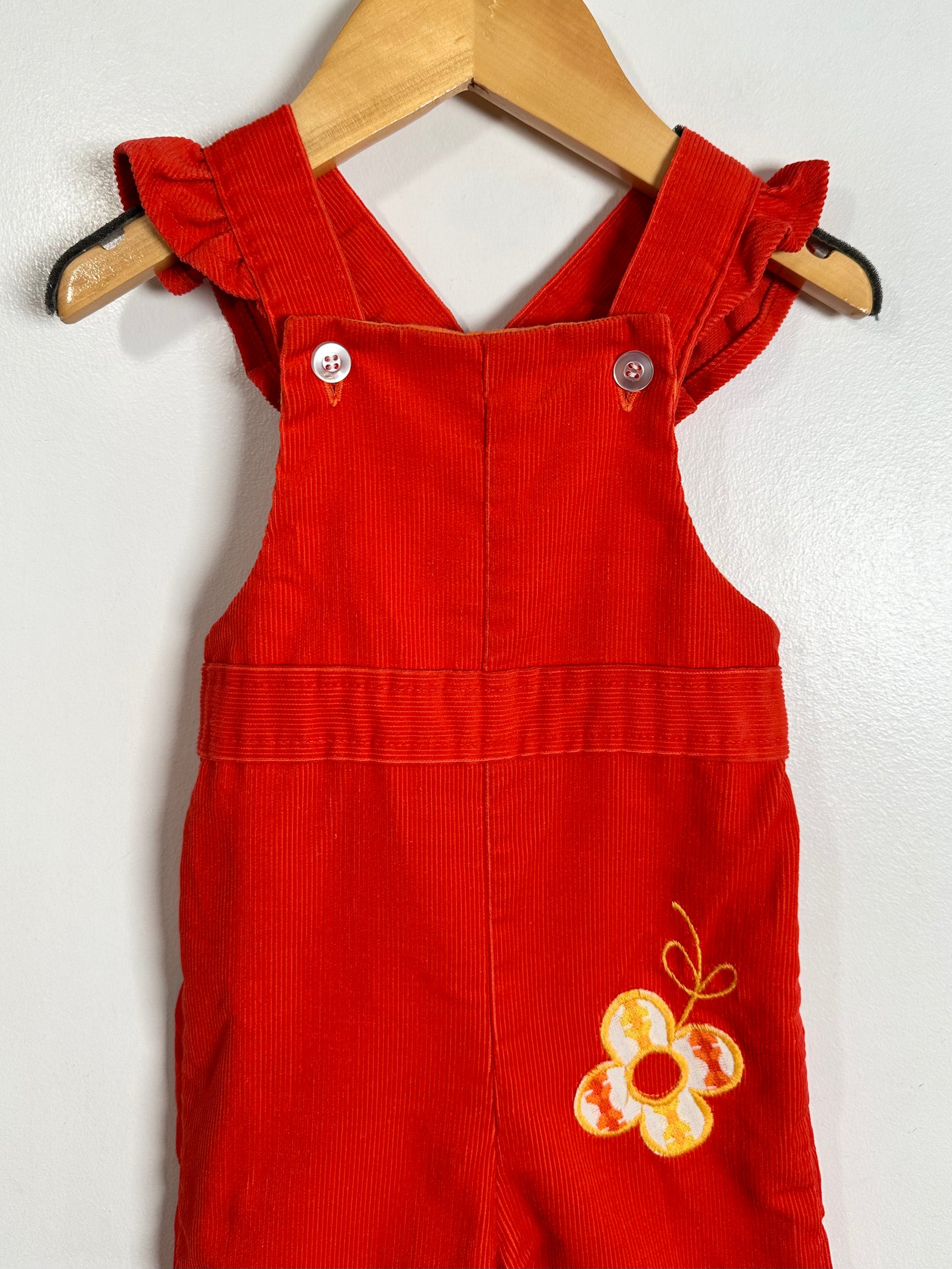 A bright red corduroy baby overall romper with ruffled shoulder straps and a yellow and white embroidered flower on the lower front, hanging on a wooden hanger against a white wall.