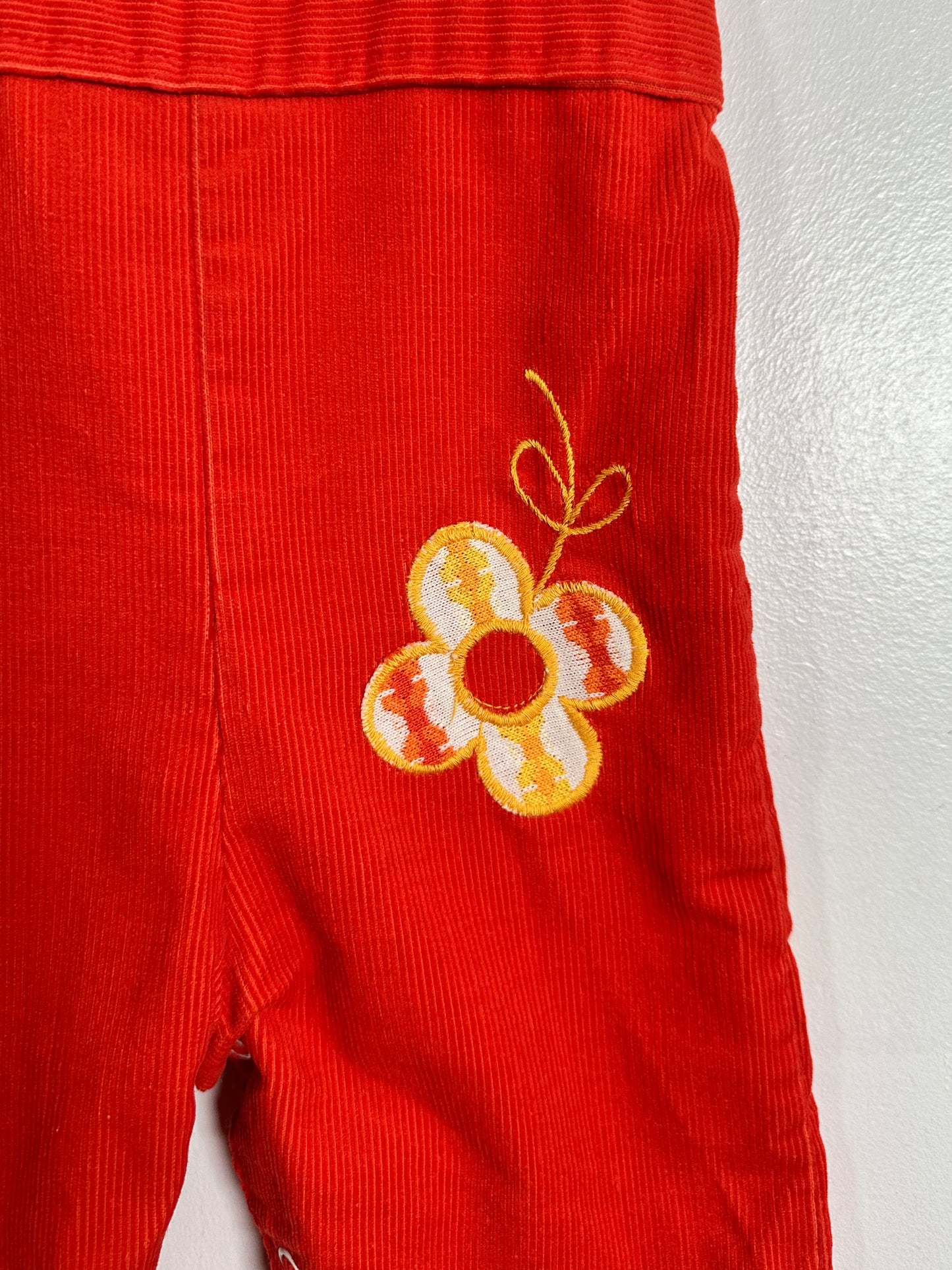 Close-up of red corduroy pants with an embroidered flower motif on the leg, featuring yellow, white, and orange details.