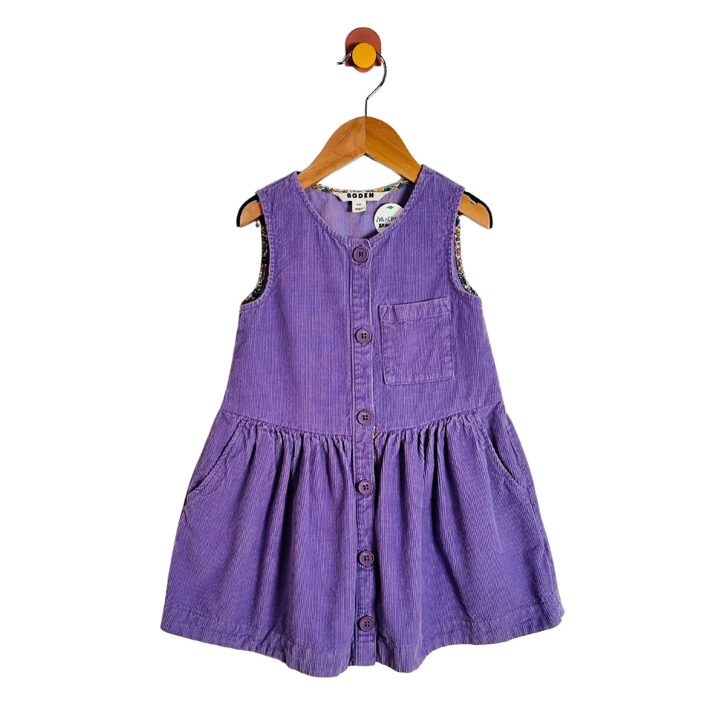 A purple corduroy sleeveless baby dress with a button-up front and chest pocket, hanging on a wooden hanger against a white background.