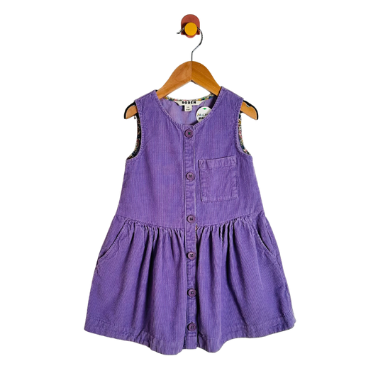 A purple corduroy sleeveless baby dress with a button-up front and chest pocket, hanging on a wooden hanger against a white background.