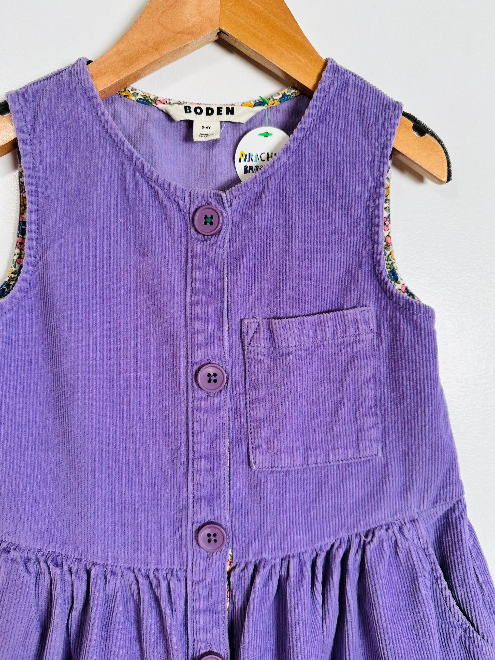 A purple corduroy sleeveless dress or pinafore top with a button front and chest pocket, hanging on a wooden hanger against a plain light background. The brand name BODEN is visible on the inner neck label.