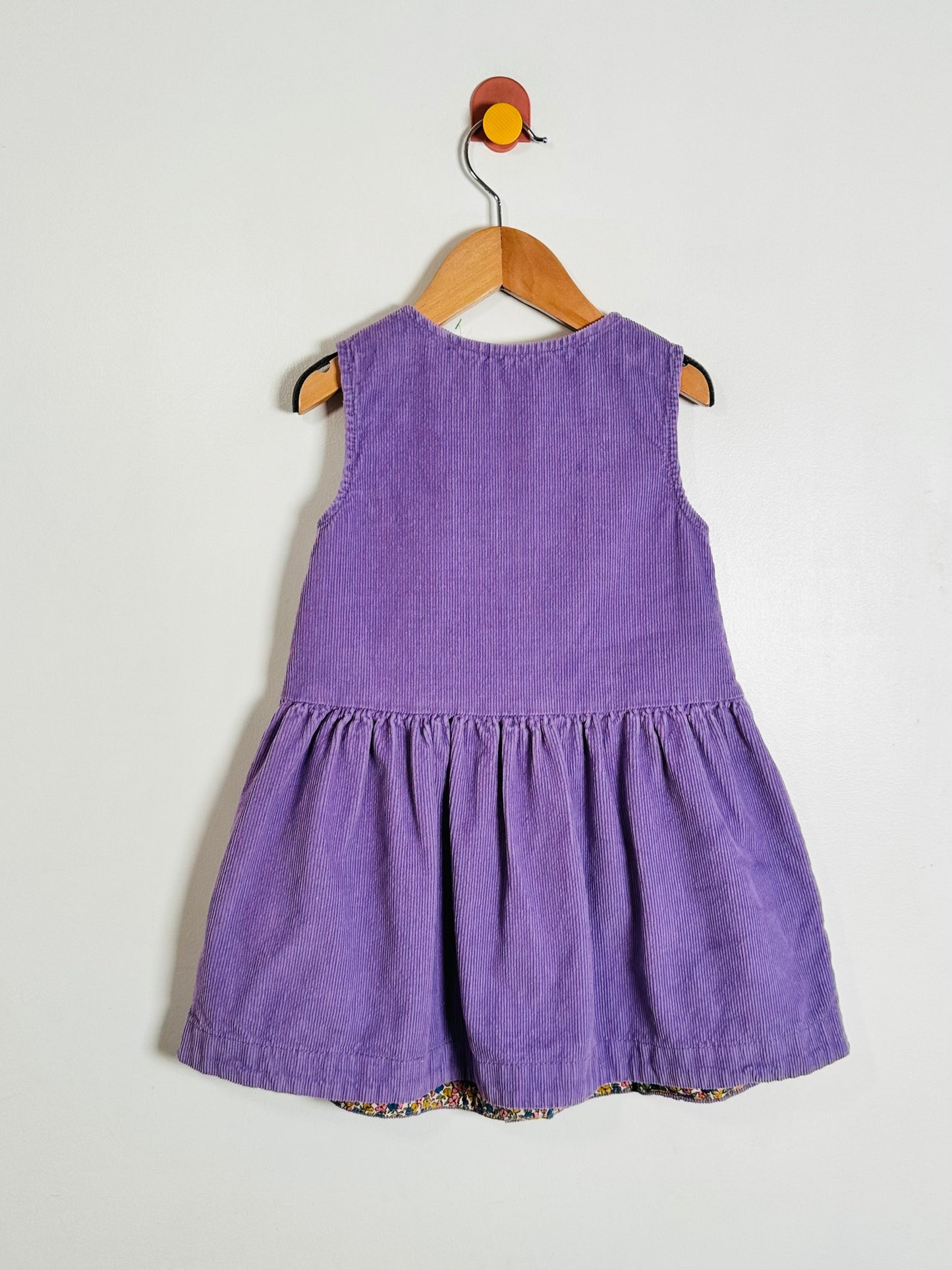 A sleeveless purple corduroy dress for a toddler, hanging on a wooden hanger against a plain off-white wall.