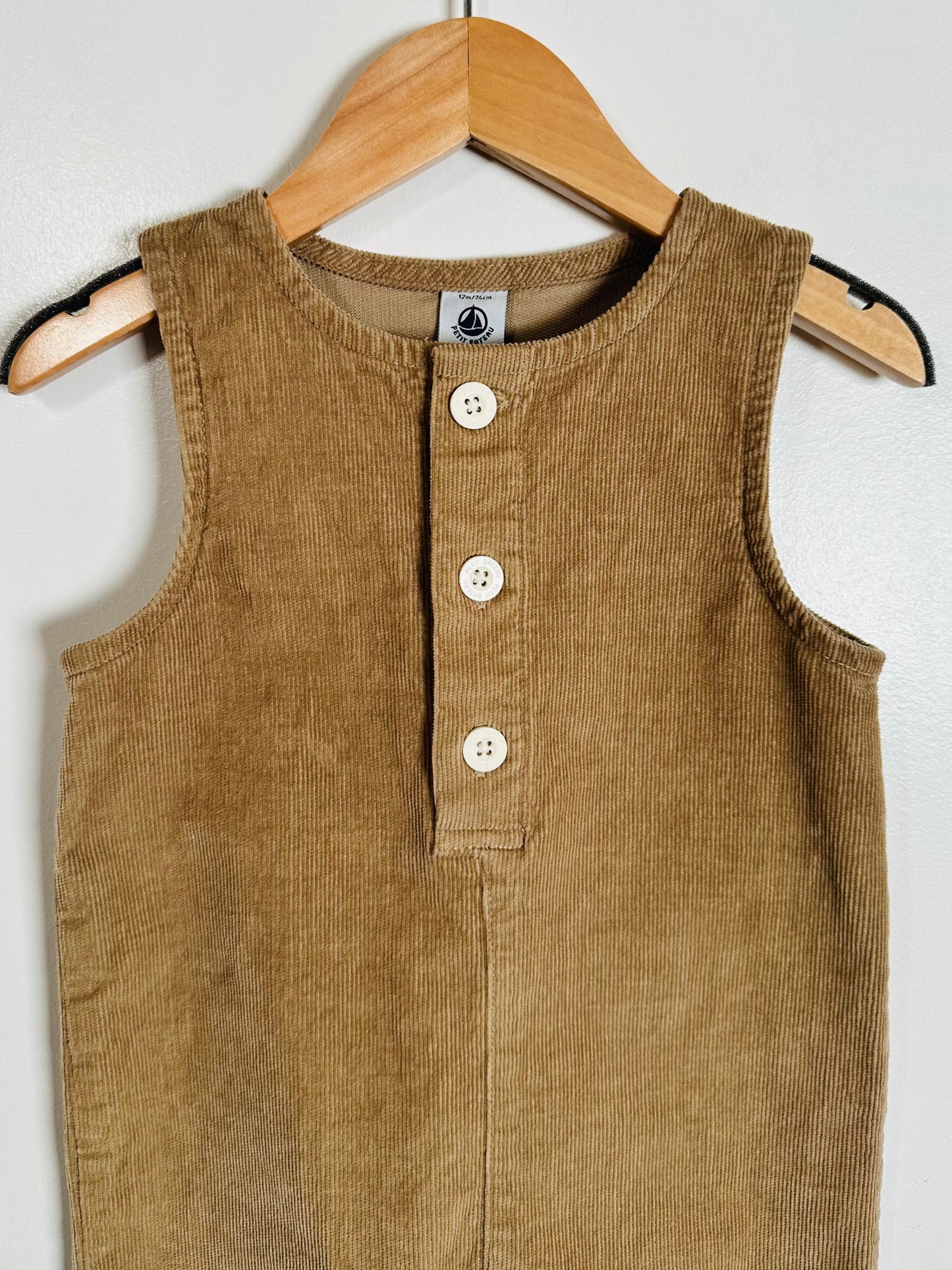 A sleeveless brown corduroy romper or long-sleeve top hanging on a wooden hanger against a white wall. It features a buttoned henley-style neckline with four white buttons.