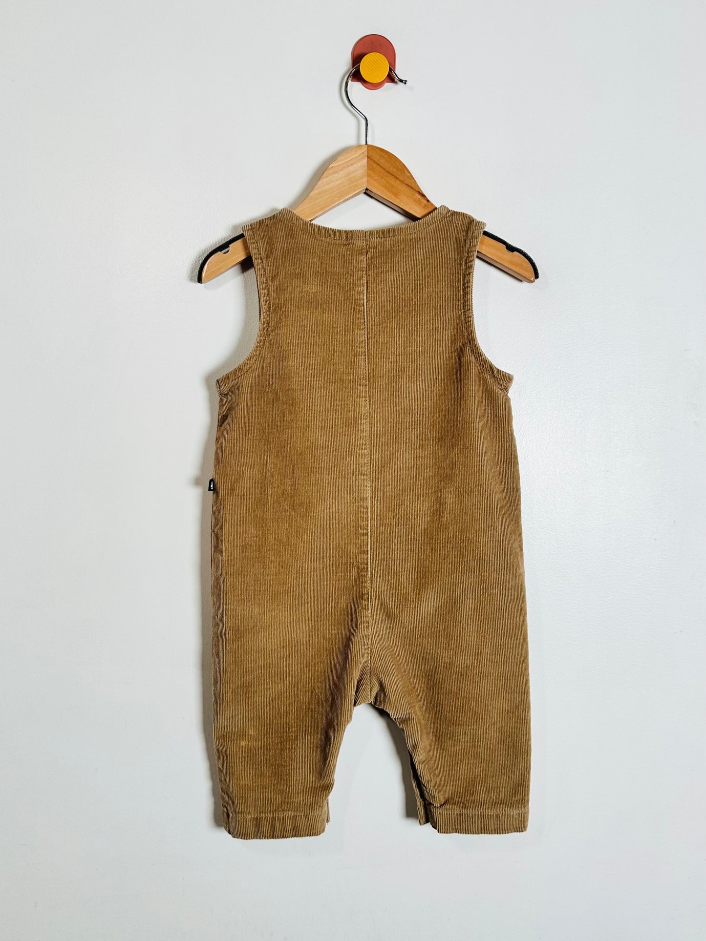 A brown corduroy baby romper hanging on a wooden hanger against a plain white wall.