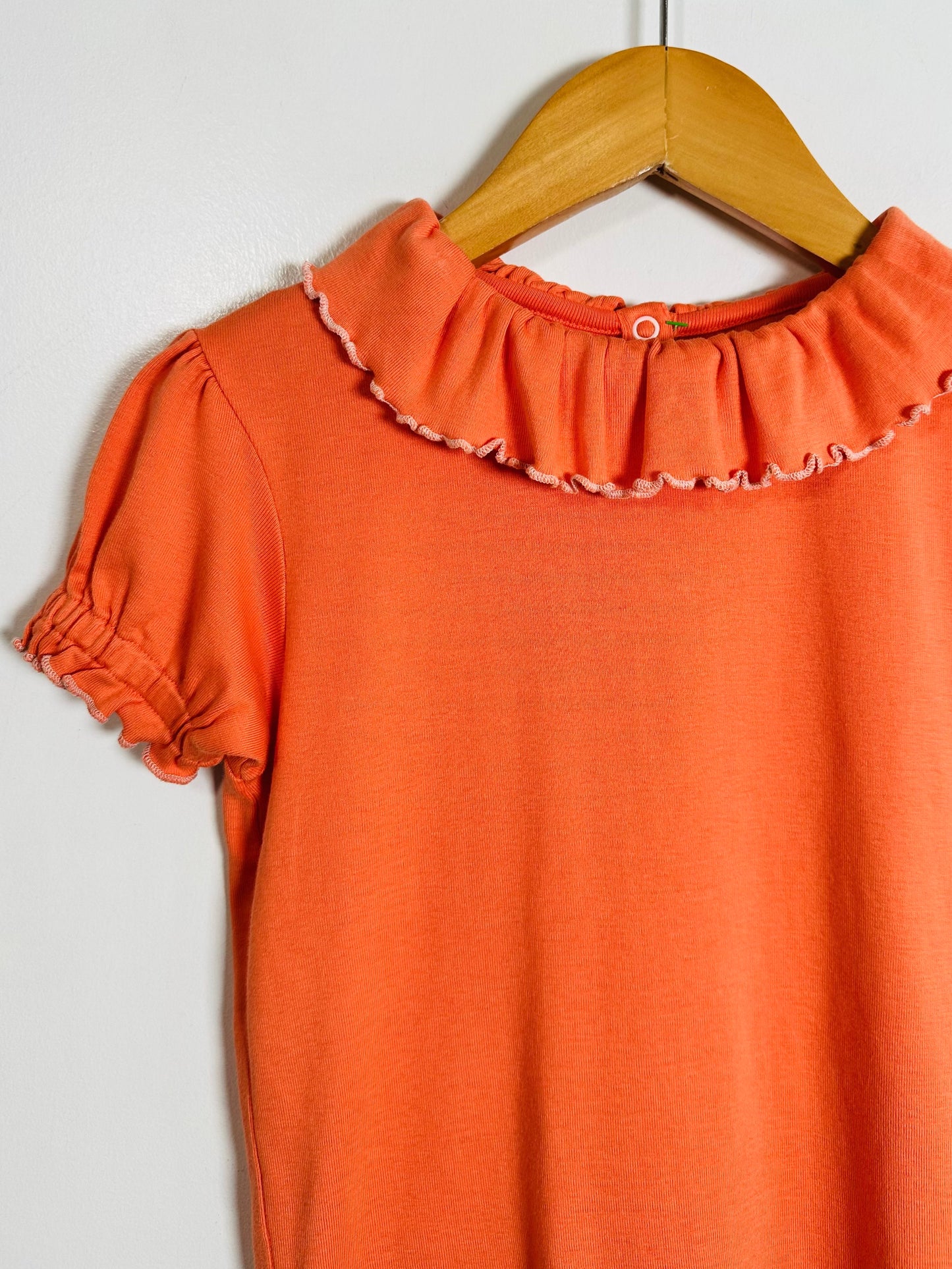 An orange short-sleeve top with a ruffled Peter Pan collar and ruffled sleeve cuffs, hanging on a wooden hanger against a plain light gray background.