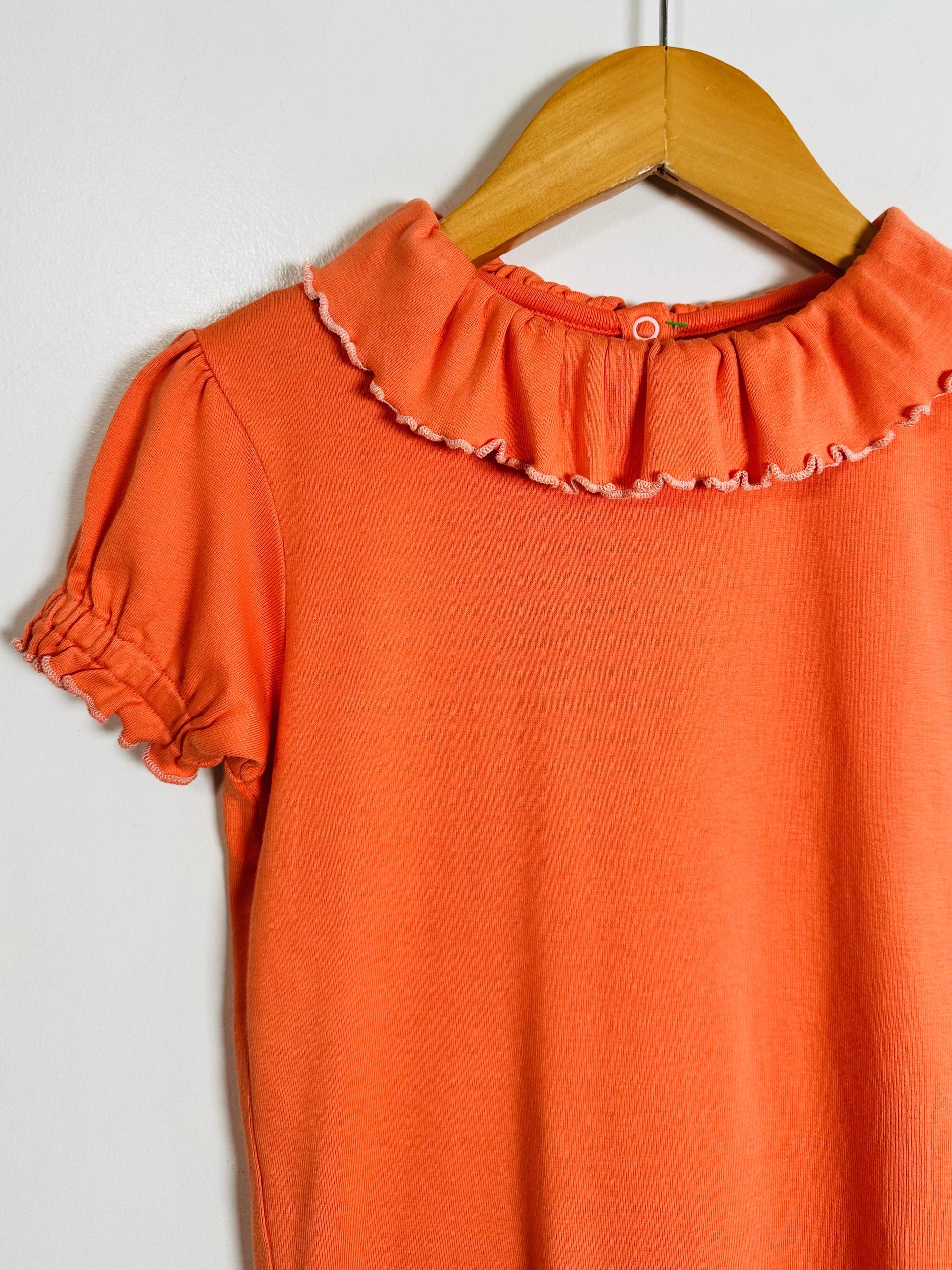 An orange short-sleeve top with a ruffled Peter Pan collar and ruffled sleeve cuffs, hanging on a wooden hanger against a plain light gray background.