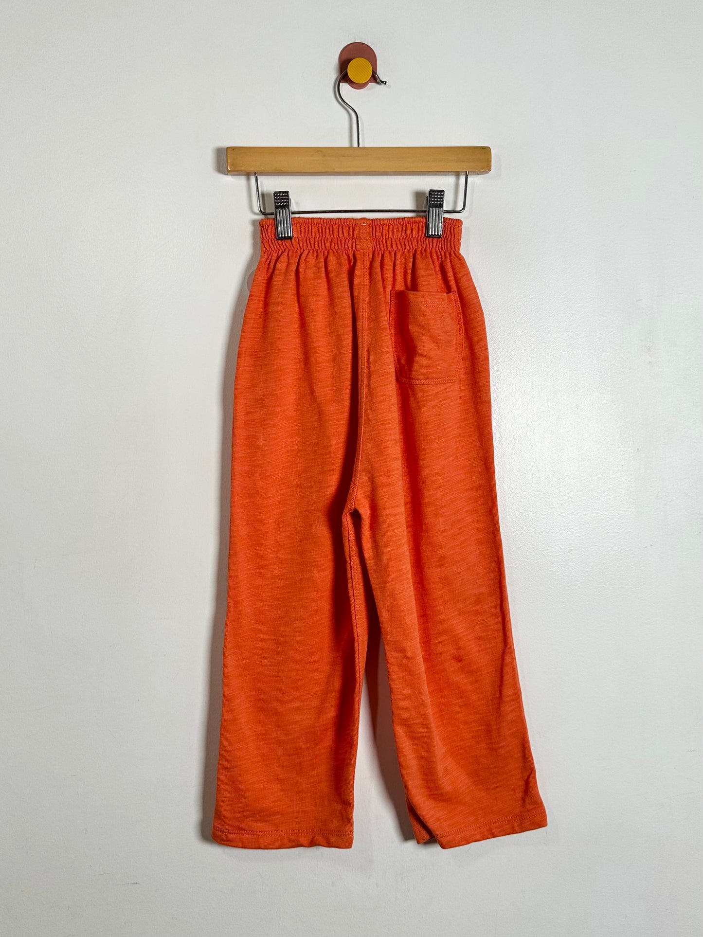 A pair of wide-leg cropped pants in a heathered burnt orange fabric, hanging from a wooden hanger against a plain off-white wall.