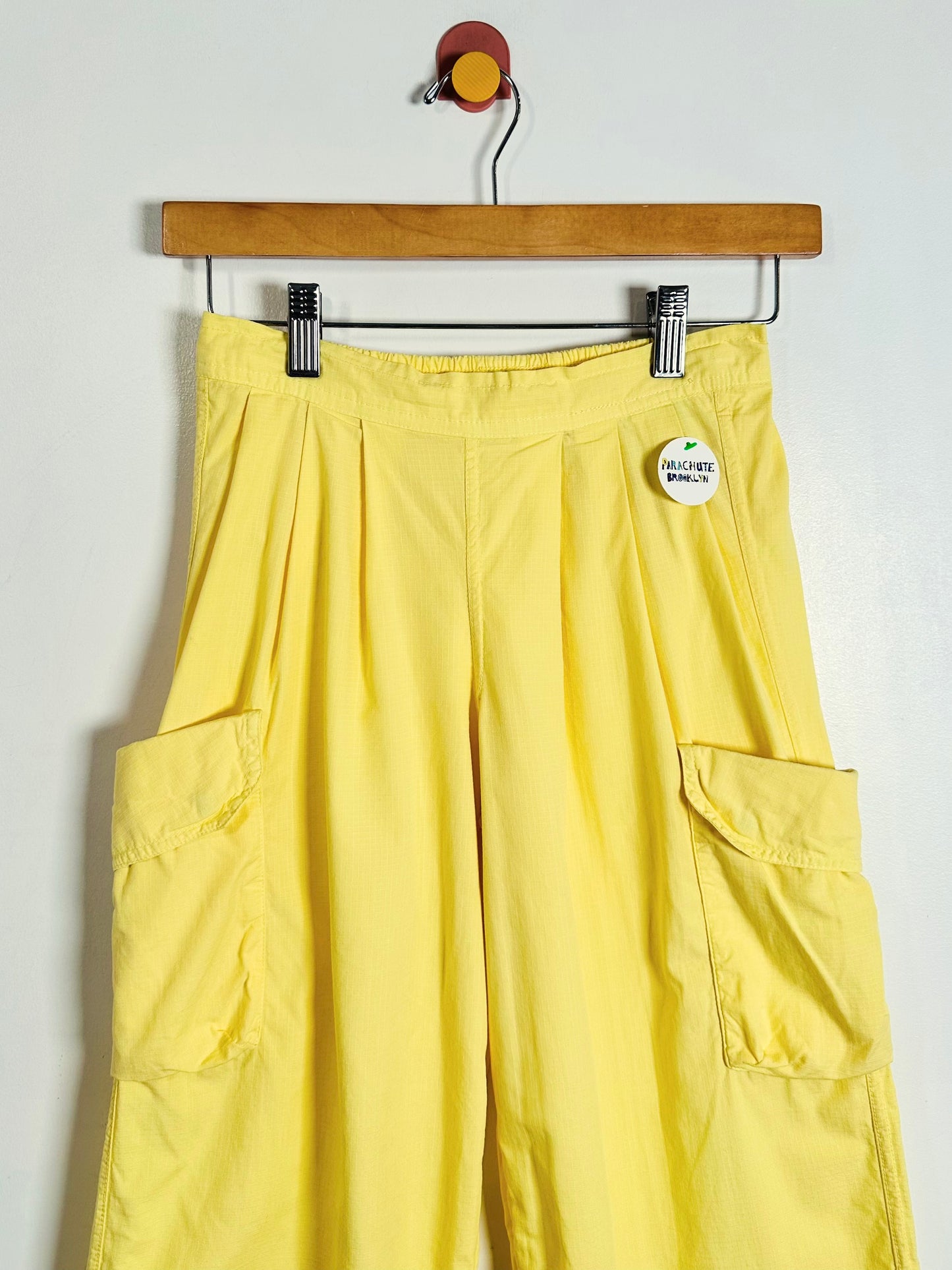 Yellow cargo pants hanging from a wooden hanger against a plain white wall, featuring side cargo pockets and a small round white tag near the waistband.