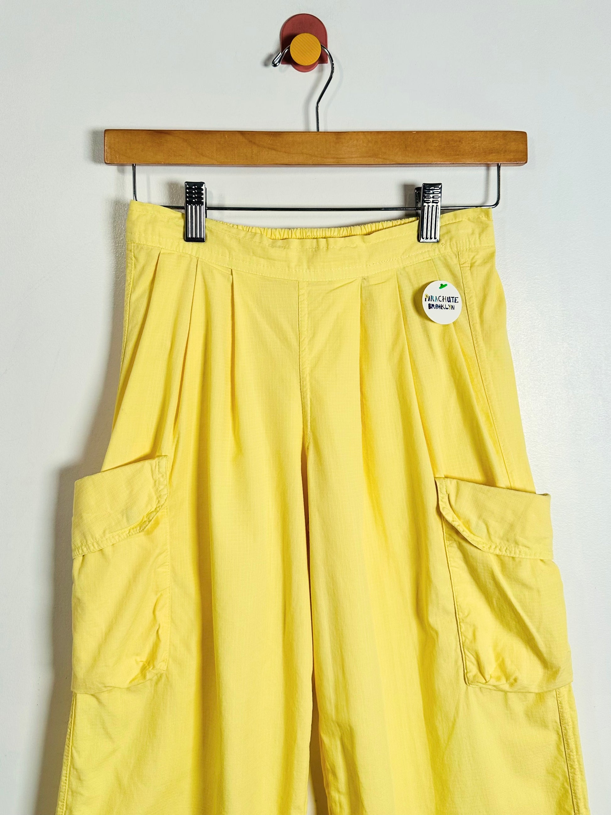 Yellow cargo pants hanging from a wooden hanger against a plain white wall, featuring side cargo pockets and a small round white tag near the waistband.