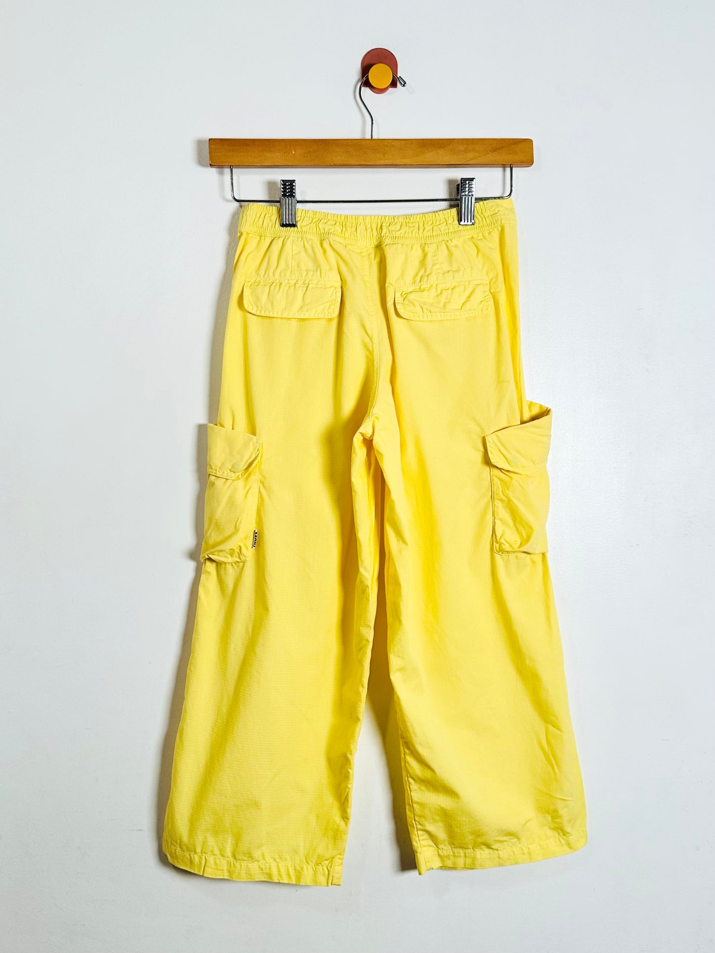 Yellow cargo pants with side flap pockets, hanging from a wooden hanger against a plain white wall.