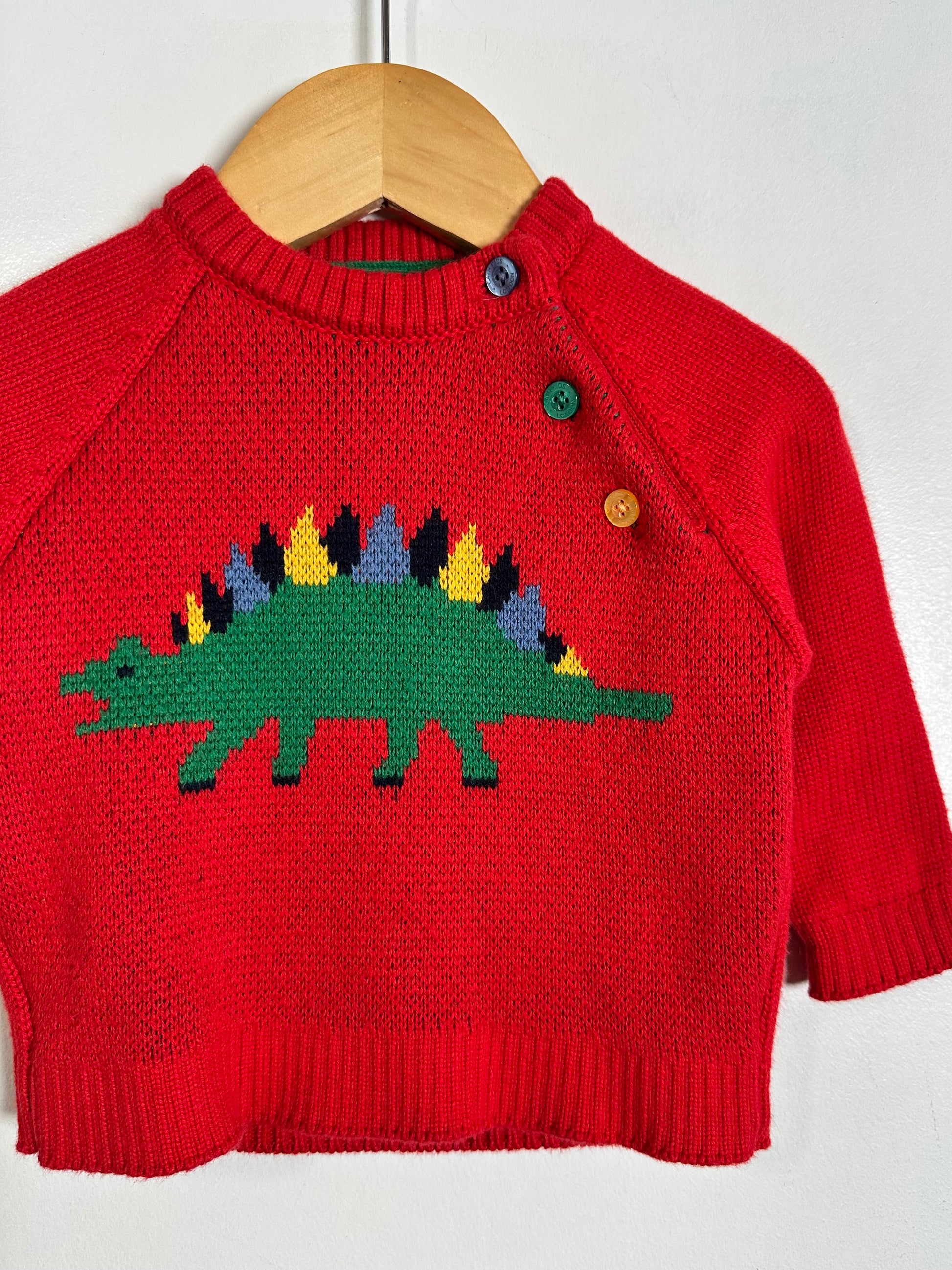 A red knit baby sweater with a colorful knitted dinosaur design on the front, hanging on a wooden hanger against a plain white wall.