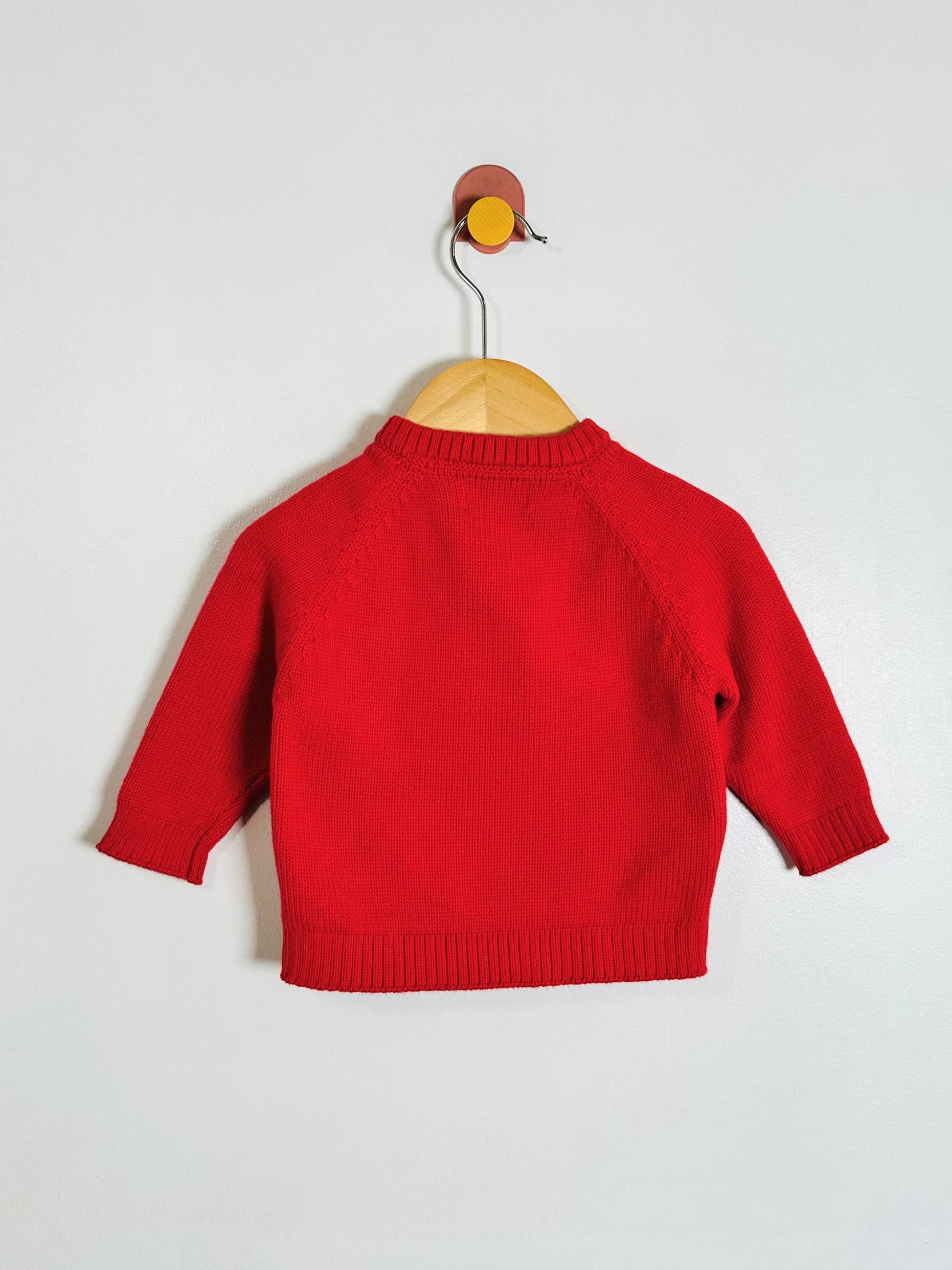 A bright red knit baby sweater, shown from the back on a wooden hanger against a plain light gray wall.
