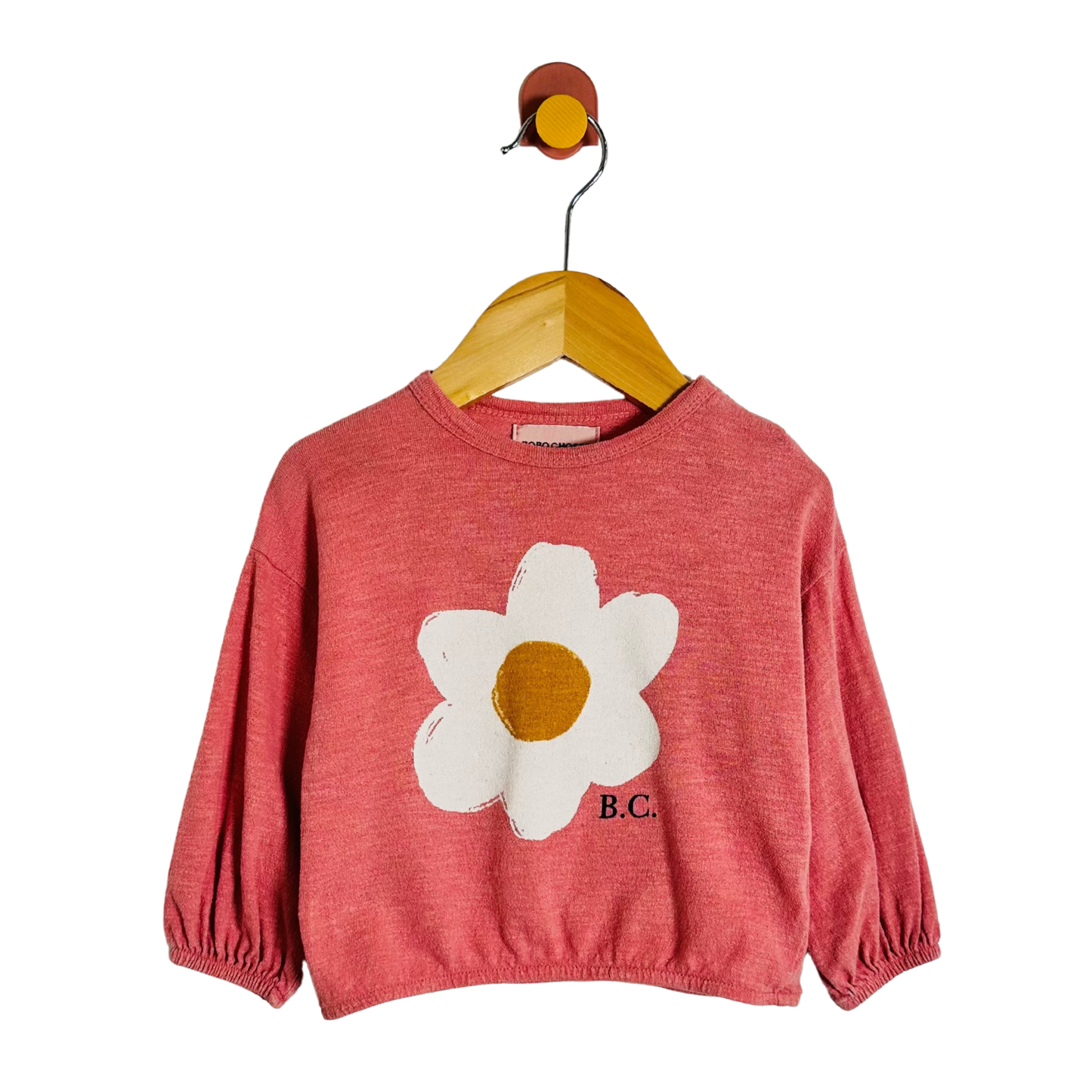 A coral pink long-sleeve crop top for kids, featuring a large white daisy graphic with a mustard yellow center on the front and 'B.C.
