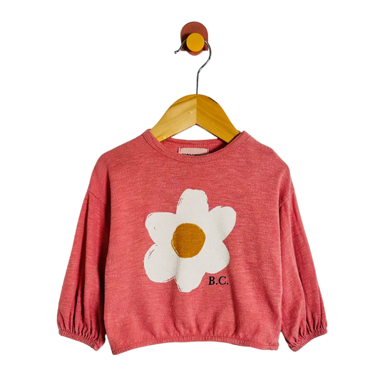 A coral pink long-sleeve crop top for kids, featuring a large white daisy graphic with a mustard yellow center on the front and 'B.C.