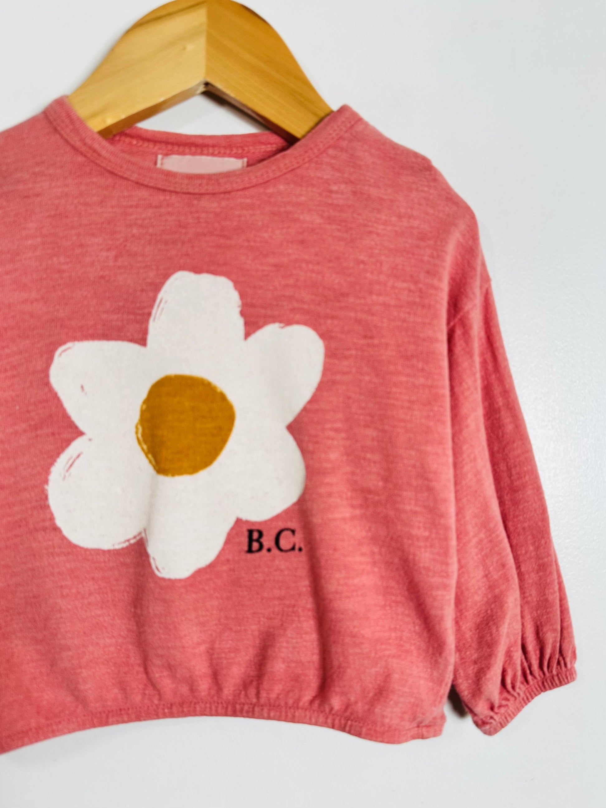 A children's long-sleeve top in heathered dusty rose pink, featuring a large white flower applique with a mustard-yellow center on the chest and the letters 'B.C.